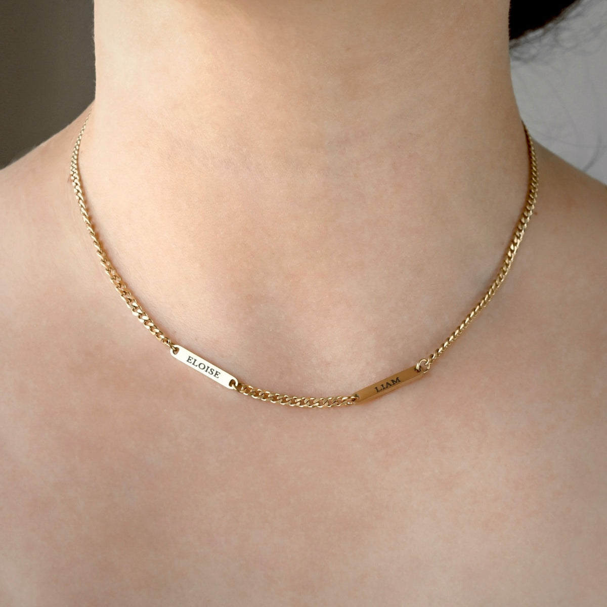 A close-up of a gold chain necklace worn on a person's neck, featuring two small engraved name plates reading 'ELOISE' and 'LIAM'.
