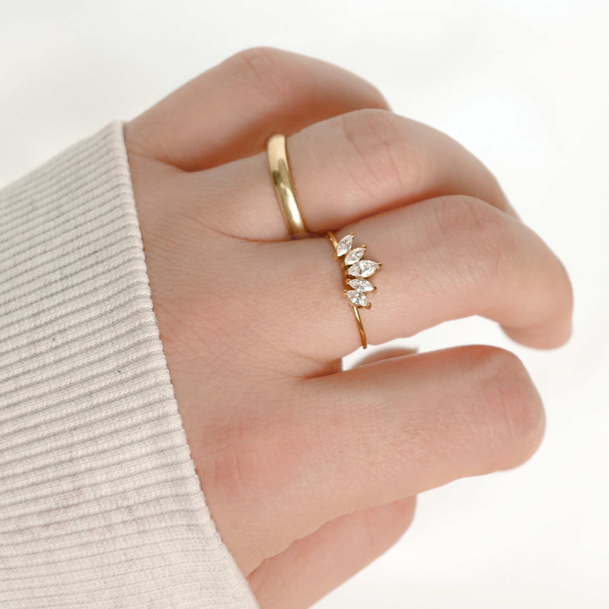 A close-up of a hand wearing two gold rings, including a delicate leaf cluster ring with small clear marquise stones on one finger, against a bright white background.