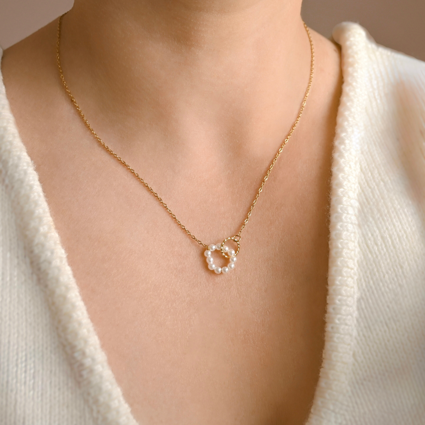 A close-up of a person wearing a delicate gold chain necklace with a small circular pendant adorned with white pearl beads and an interlocking small twisted circle, paired with a cream-colored knit top.