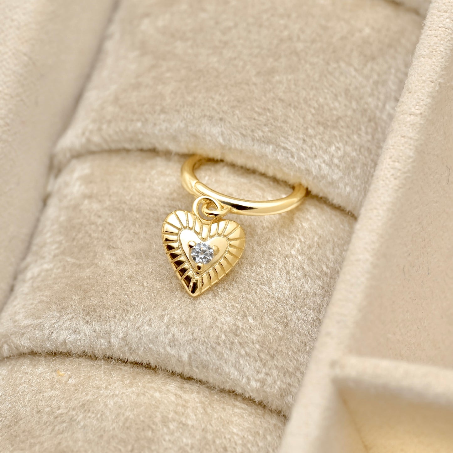 A gold heart-shaped charm hoop earrings with a small clear gemstone in the center, displayed on a beige velvet jewelry box.