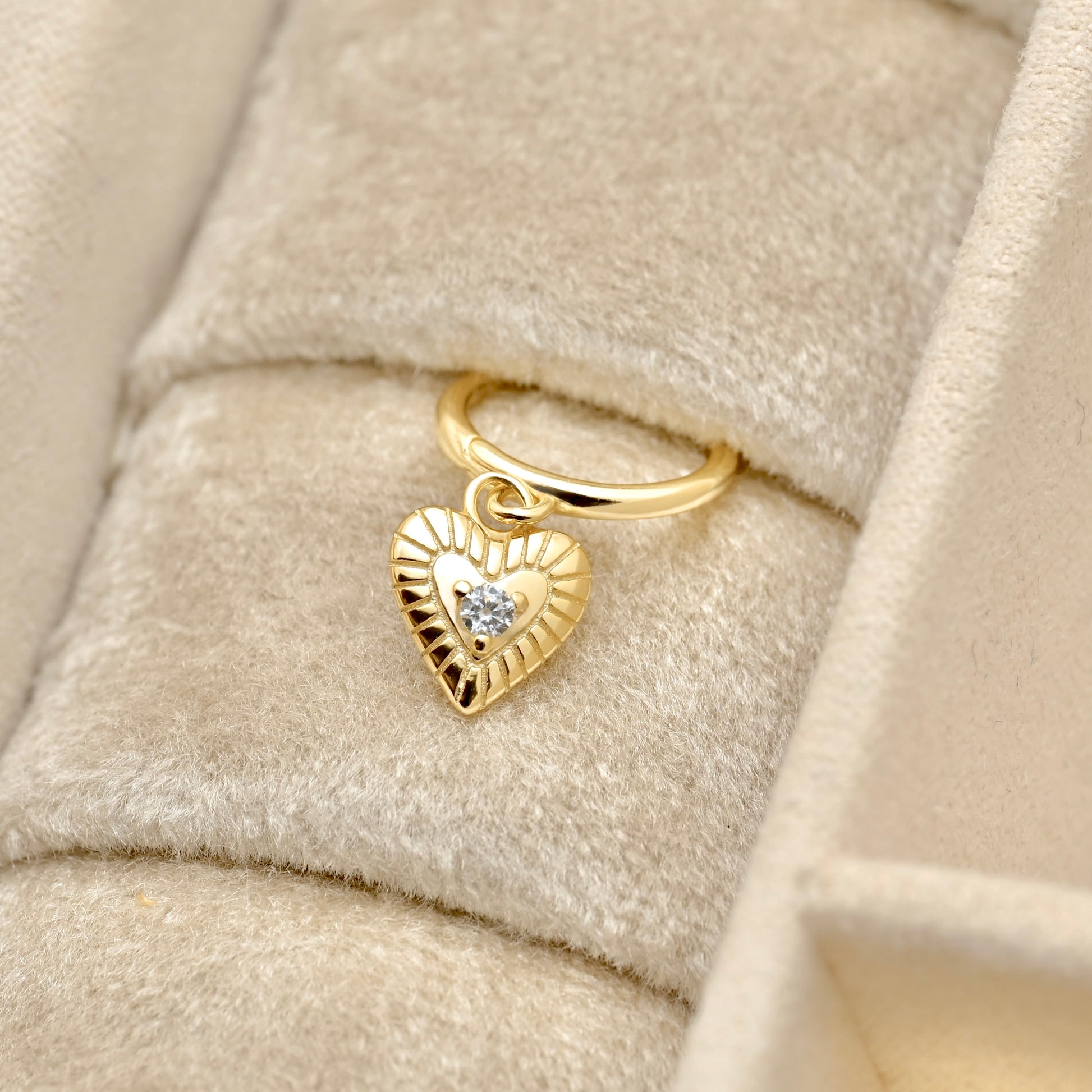 A gold heart-shaped charm hoop earrings with a small clear gemstone in the center, displayed on a beige velvet jewelry box.