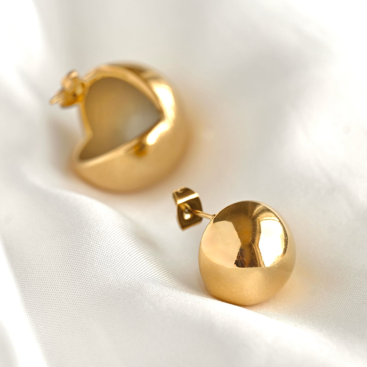 A pair of polished gold dome stud earrings resting on a soft, draped white fabric background.