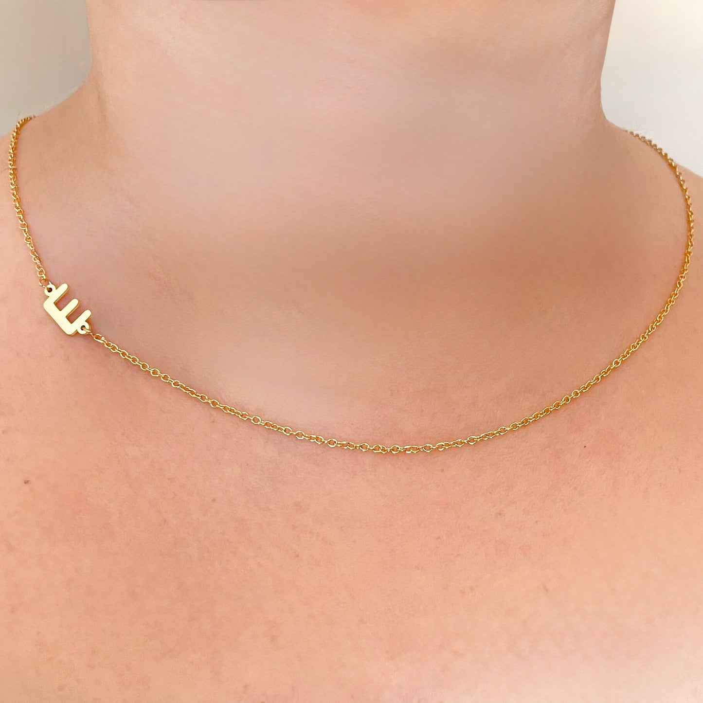 Gold necklace with a small initial positioned sideways on the side  on a person's neck against a neutral background