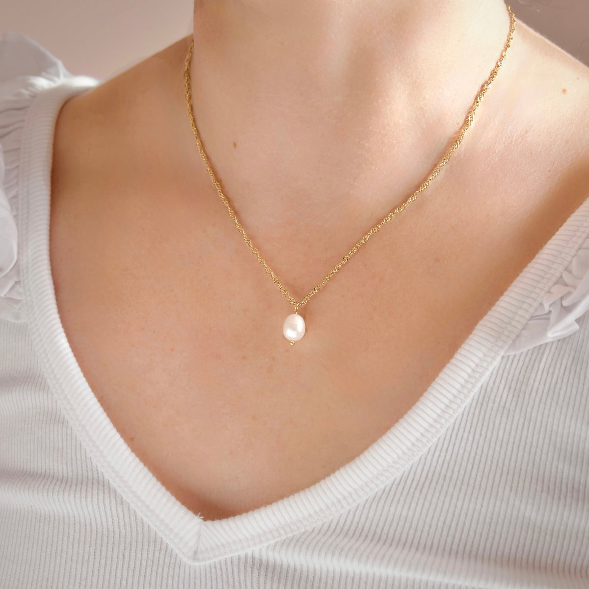 A close-up of a person wearing a delicate gold chain necklace with a single white pearl pendant, styled with a white ribbed V-neck top.