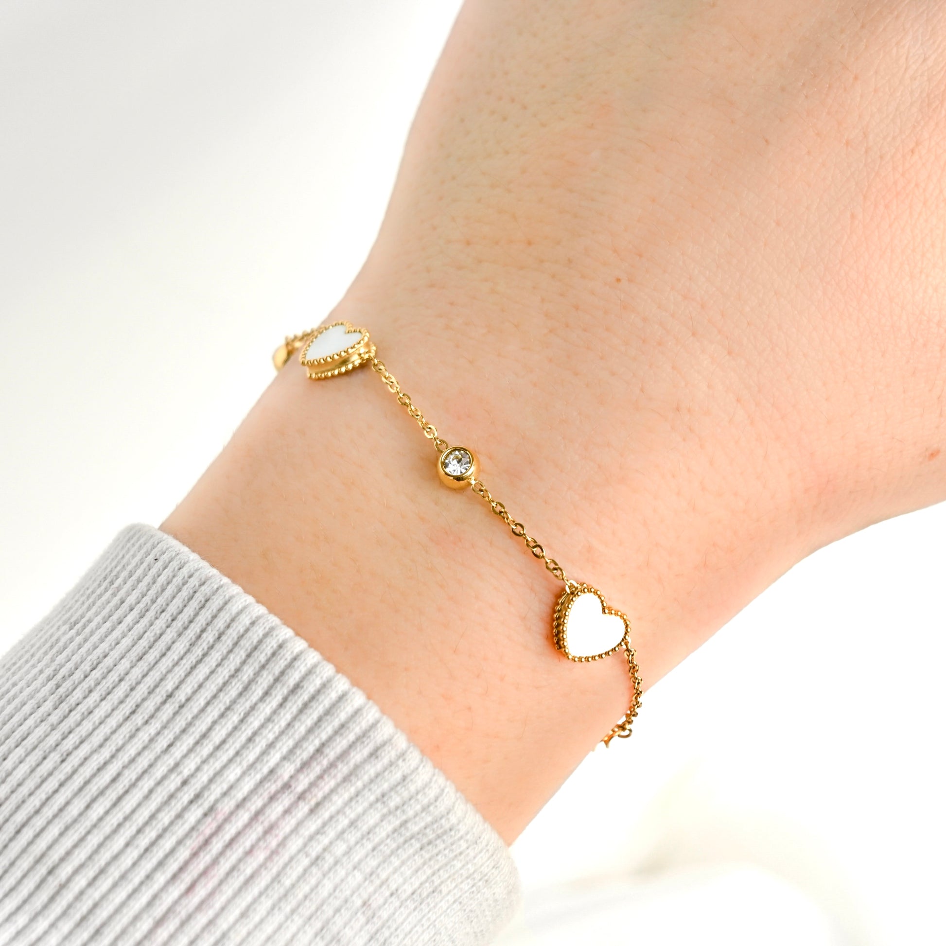 A delicate gold chain bracelet with two white heart charms and a small clear stone, shown worn on a person's wrist.