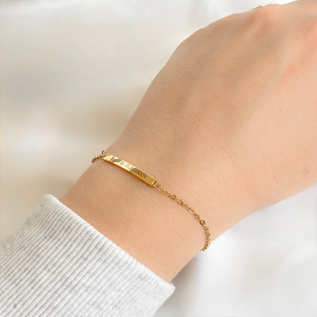 A delicate gold bar bracelet engraved with the name 'Emily' and roman numerals in a person's wrist, shown wearing a light gray ribbed knit sweater cuff against a soft white background.