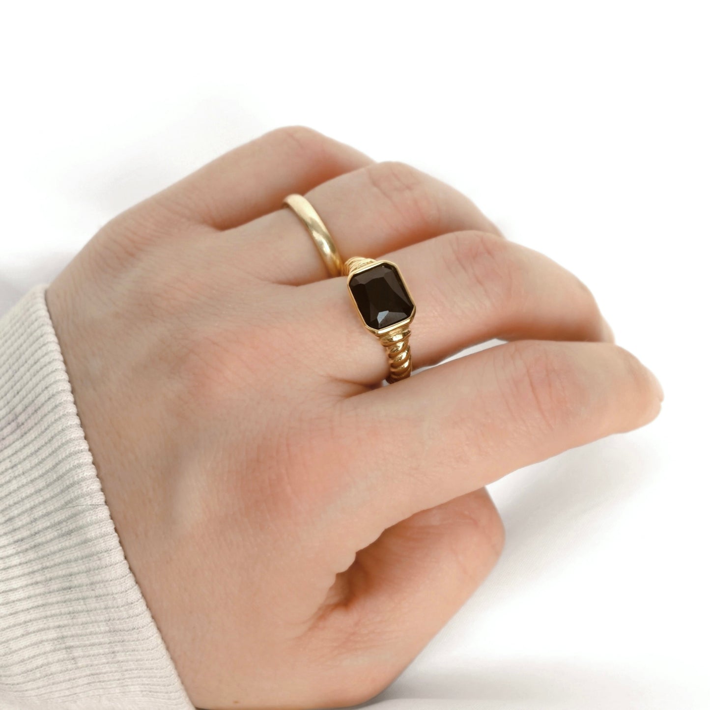 A gold ring with a rectangular dark black gemstone, worn on a person's finger against a clean white background.