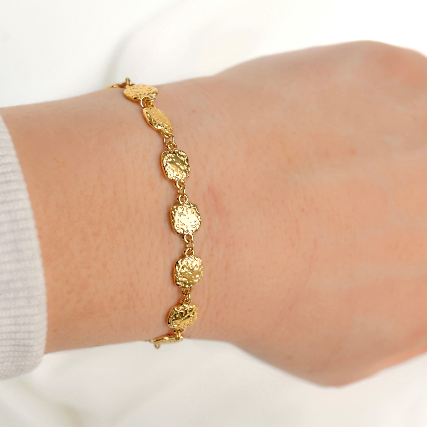 A delicate gold chain bracelet with textured, coin-like oval links worn on a person's wrist, shown against a soft white background.