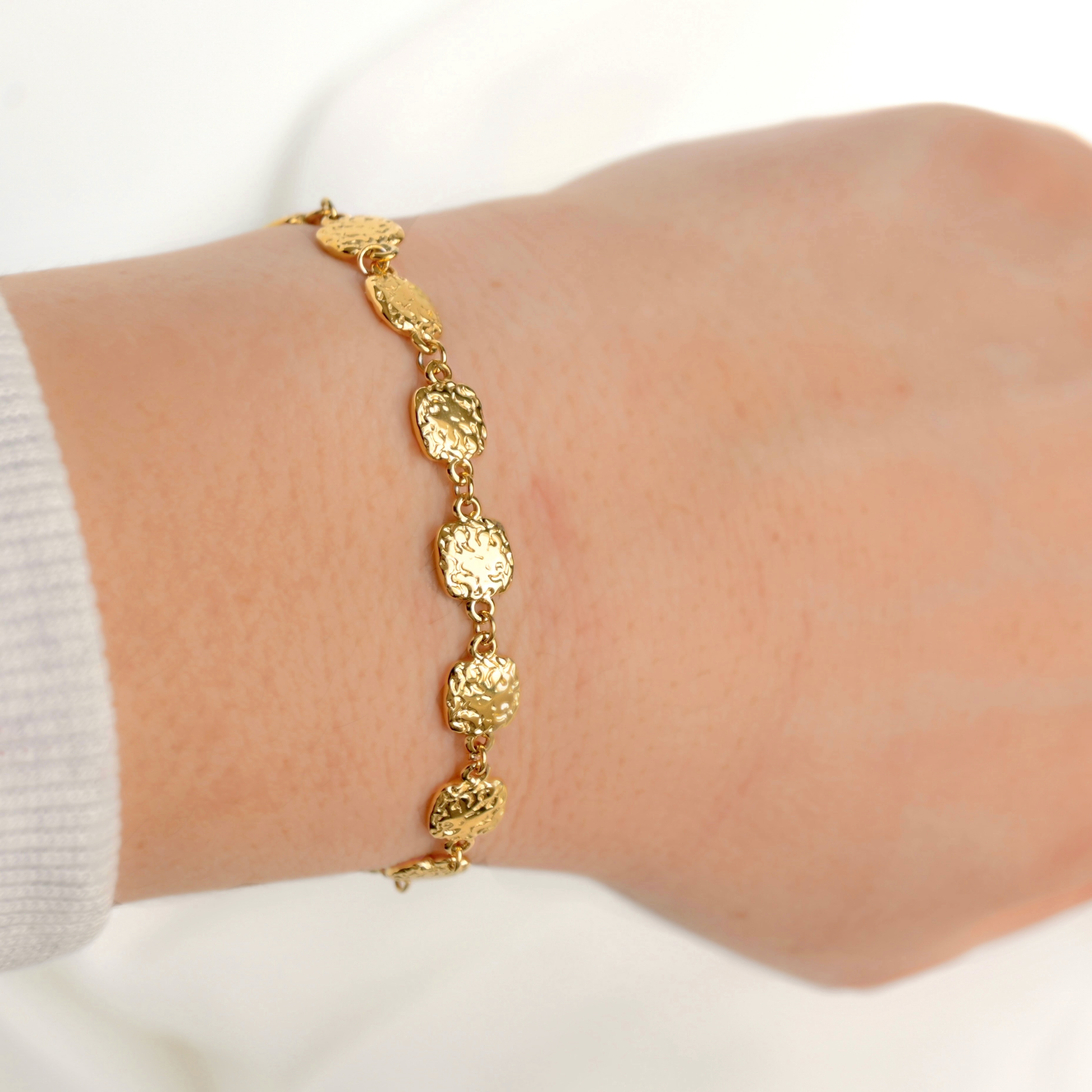 A delicate gold chain bracelet with textured, coin-like oval links worn on a person's wrist, shown against a soft white background.