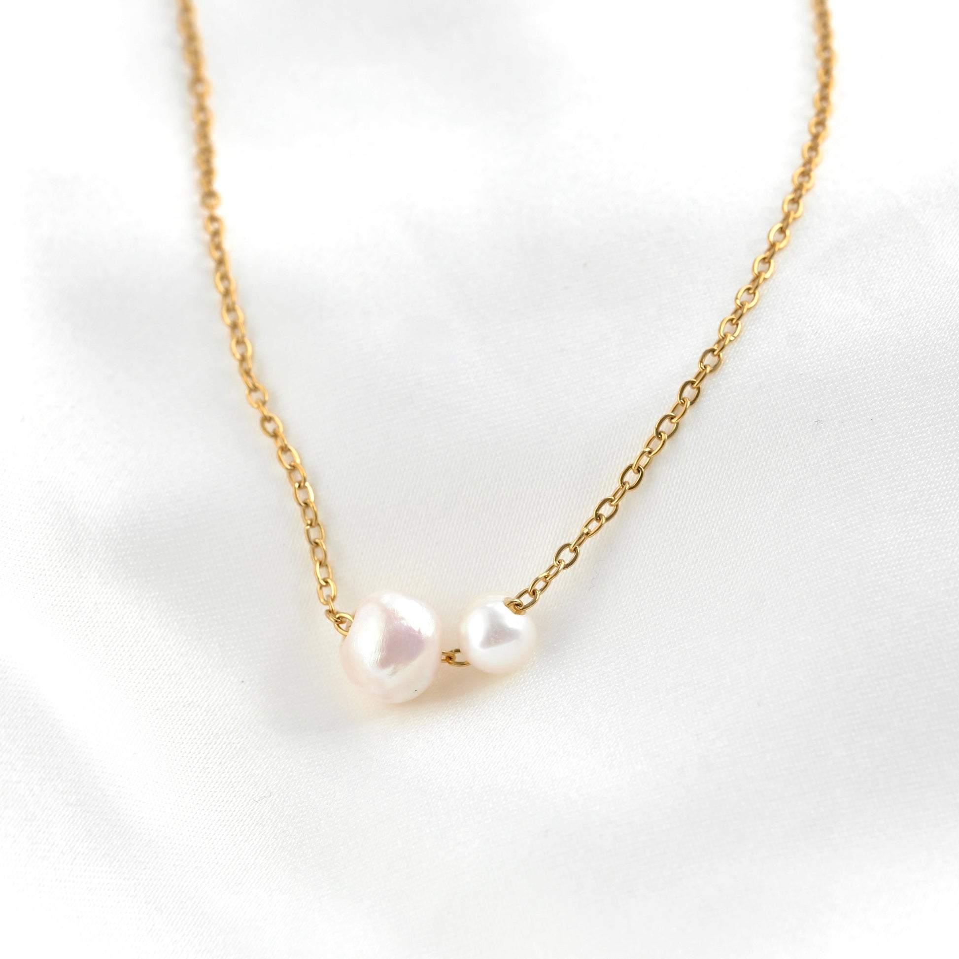A delicate gold chain necklace featuring two irregular, lustrous white freshwater pearls at the center on a thin cable chain, displayed on a soft white fabric background.