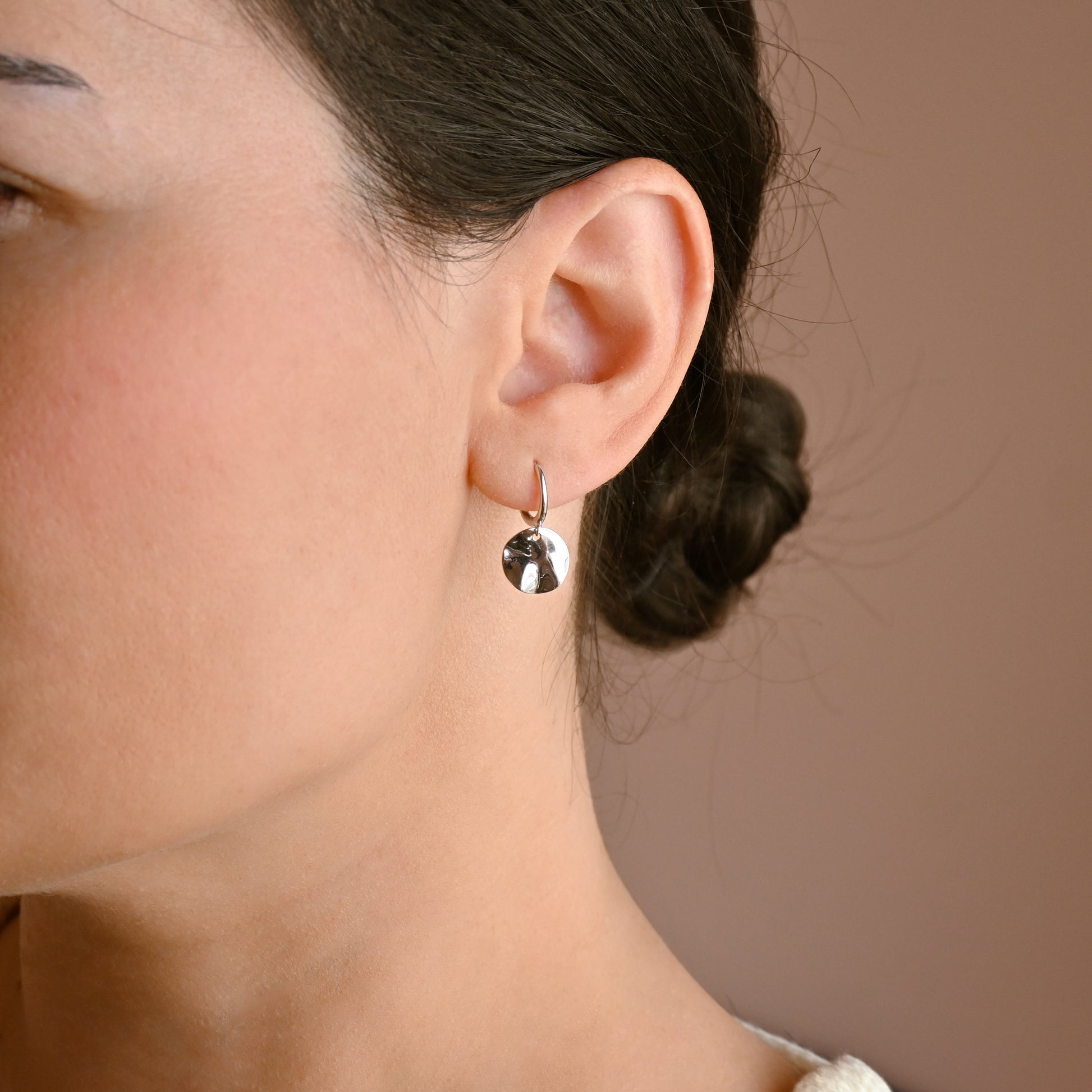 A close-up side profile of a woman wearing a small, polished silver hoop earring with a dangling  hammered spherical disc , set against a solid warm brown background.