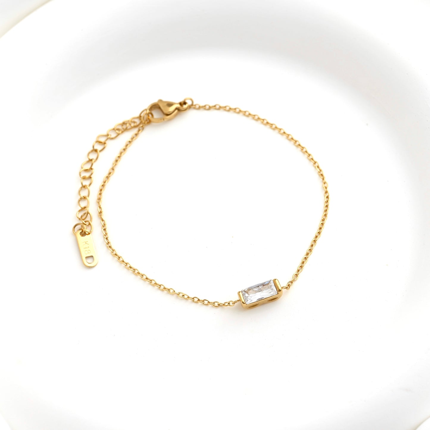 A delicate gold chain bracelet with a small baguette cut CZ stone centerpiece, displayed on a clean white background.