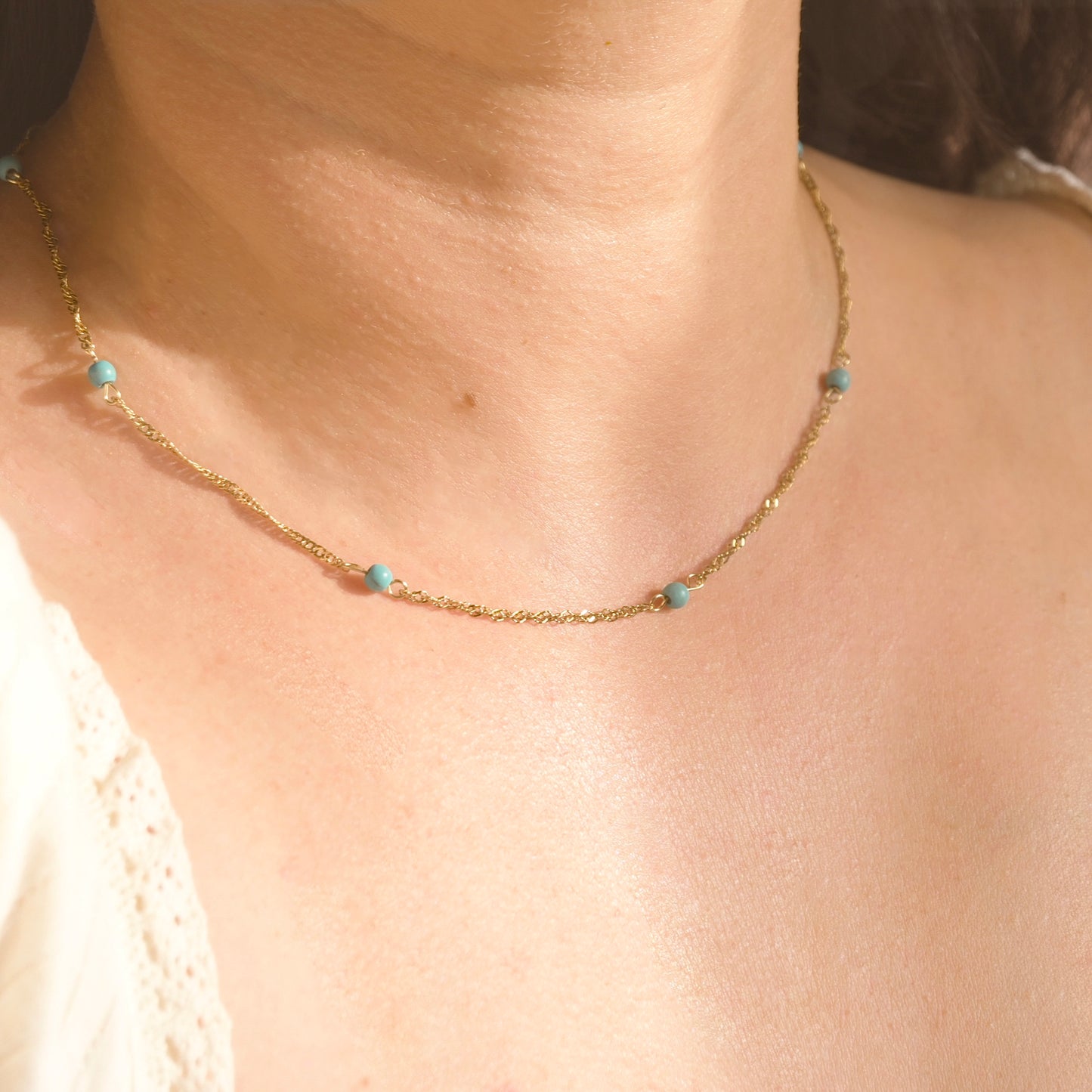 A close-up of a person's neck and chest wearing a delicate gold chain necklace with small turquoise blue beads spaced along the chain.