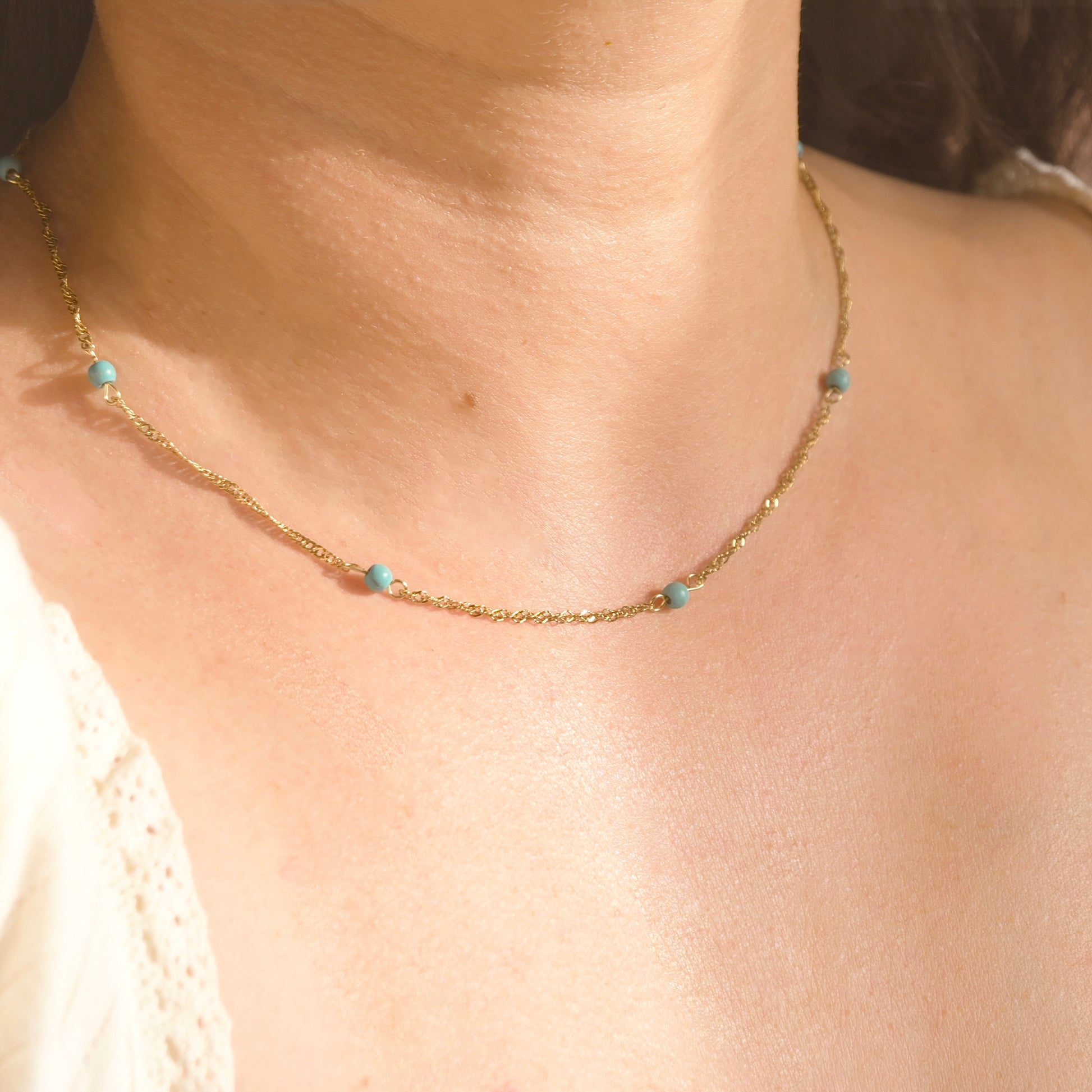 A close-up of a person's neck and chest wearing a delicate gold chain necklace with small turquoise blue beads spaced along the chain.