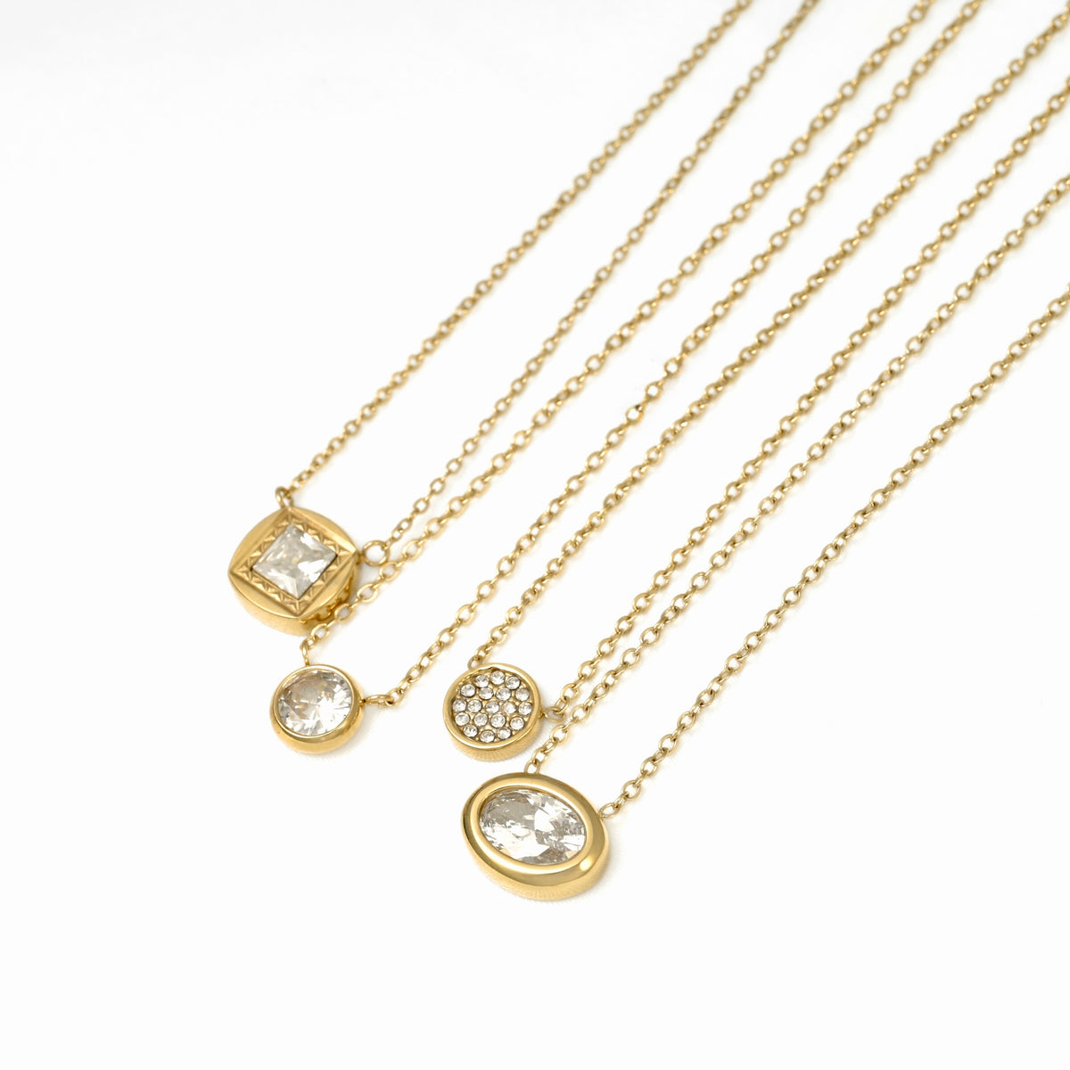 Four gold necklaces with different pendant designs, including a square bezel-set clear stone, a round pavé clear stone, and an oval clear stone, arranged on a white background.