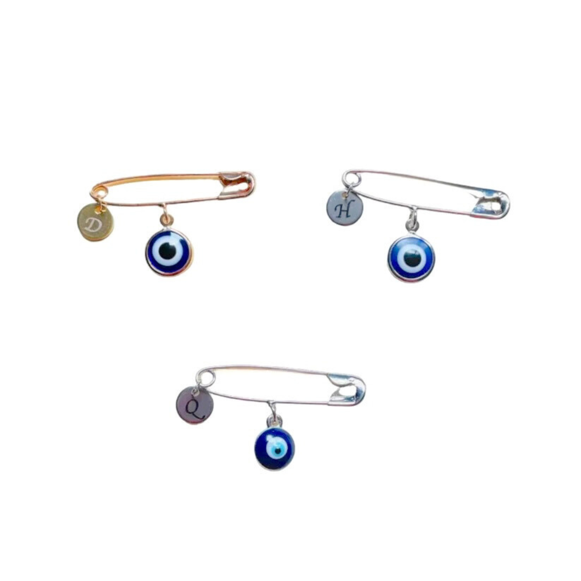 Three decorative brooches with blue eye designs and initials on a white background