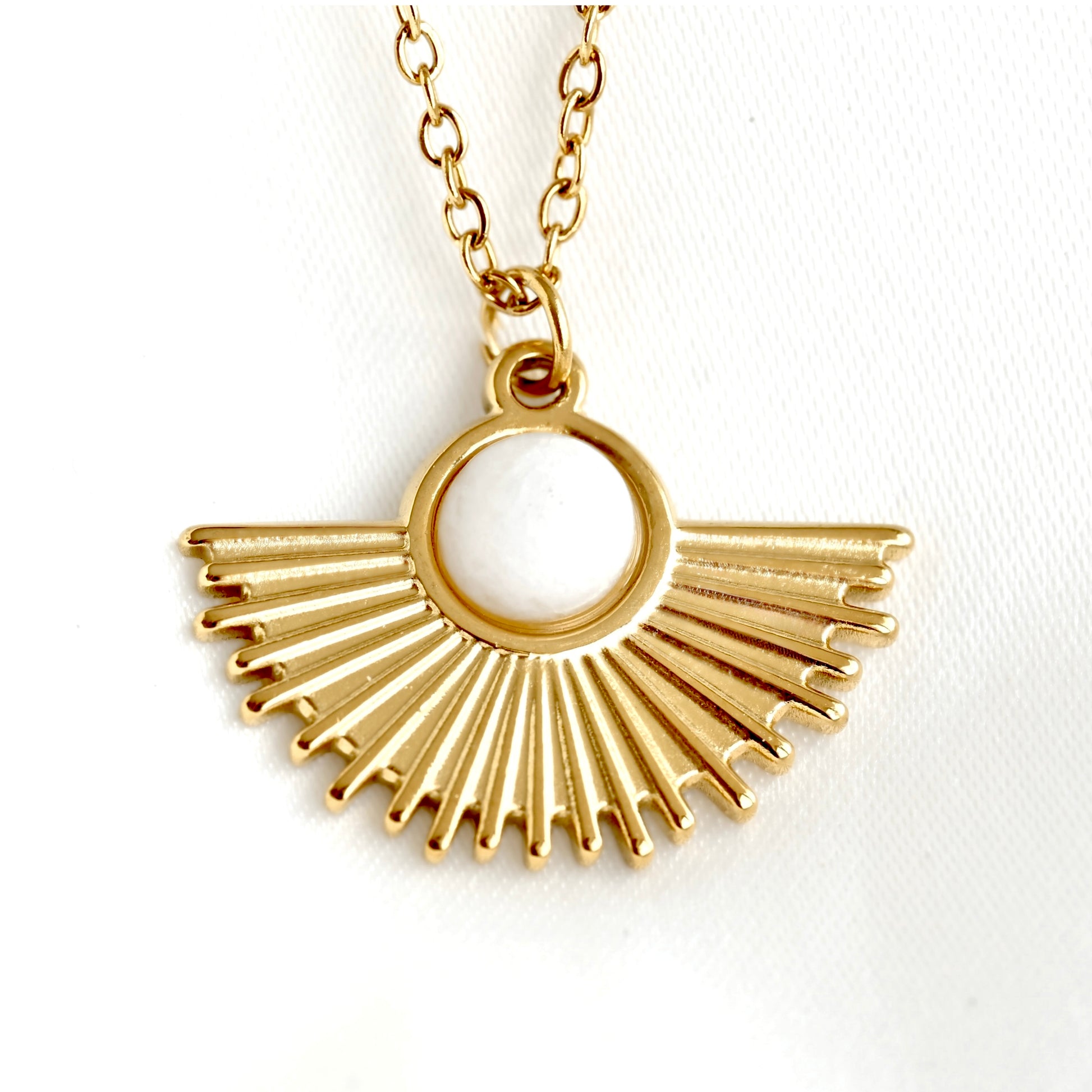 Front close up view of a gold fan pendant necklace featuring a sunburst fan design with a central white round stone, shown on a white textured fabric background.