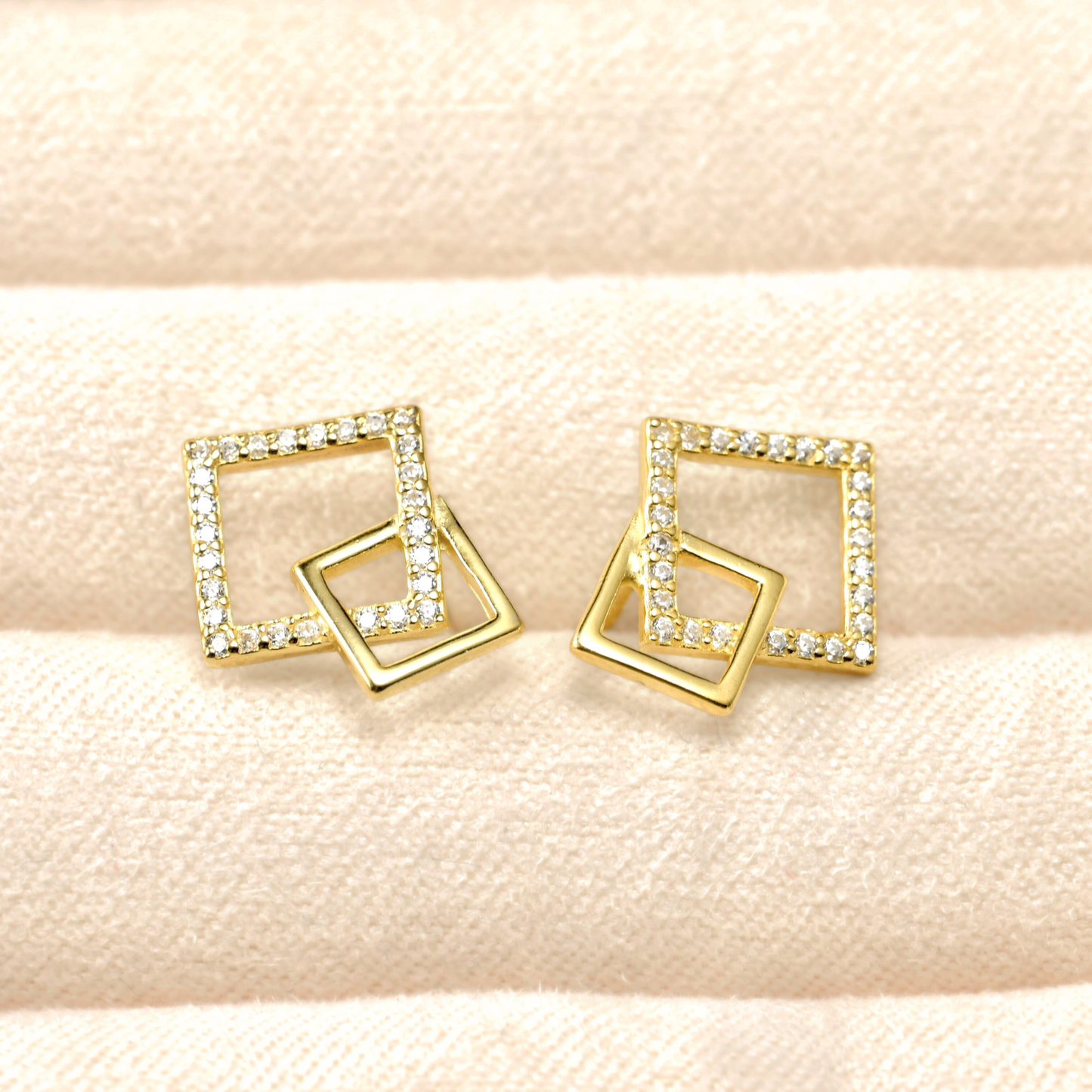 Gold square-shaped earrings with clear stones on a beige fabric background