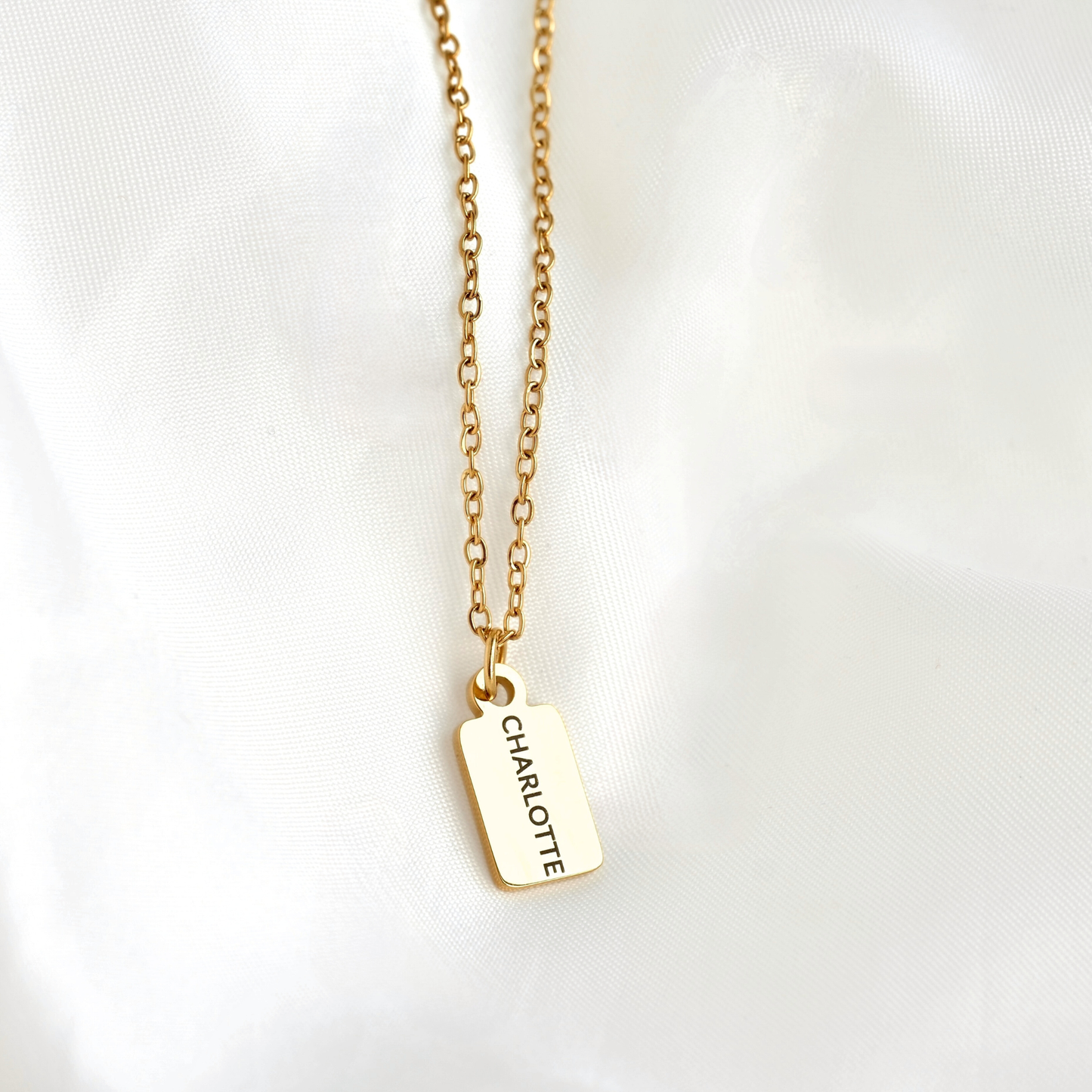 A gold chain necklace with a rectangular gold tag pendant engraved with the name 'CHARLOTTE', displayed on a soft white fabric background.