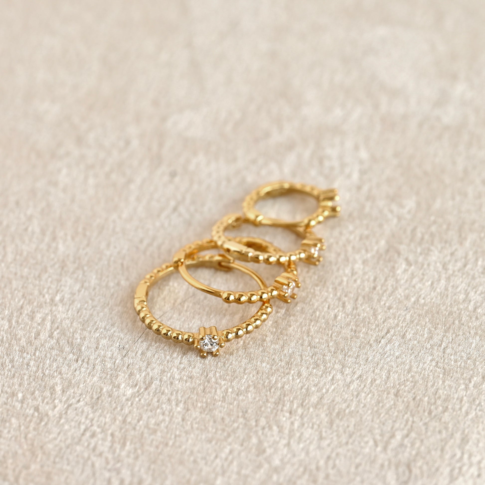 Four gold hoop earrings with textured beaded edges and small clear gemstone accents, arranged on a soft beige fabric background.