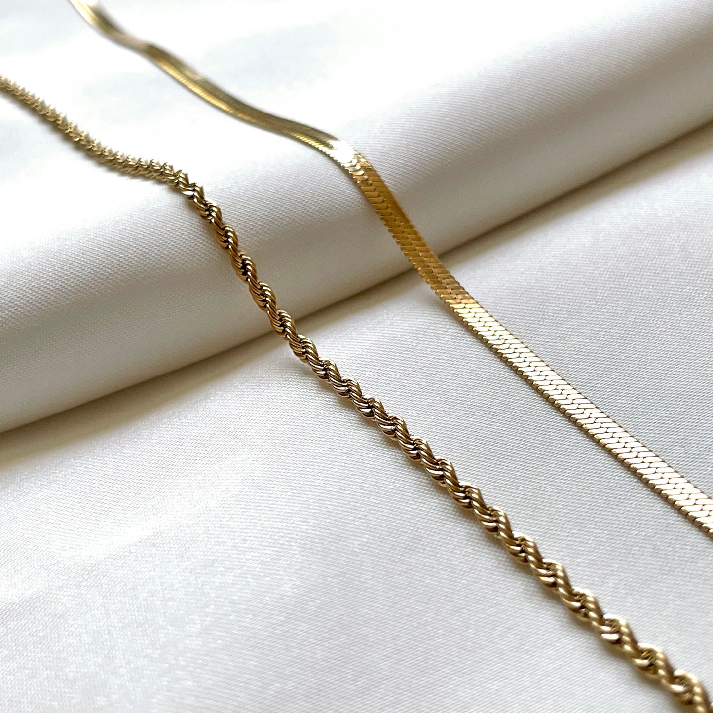 Close up of two gold chains , one 3mm herringbone and one 2mm rope chain, on a white fabric background
