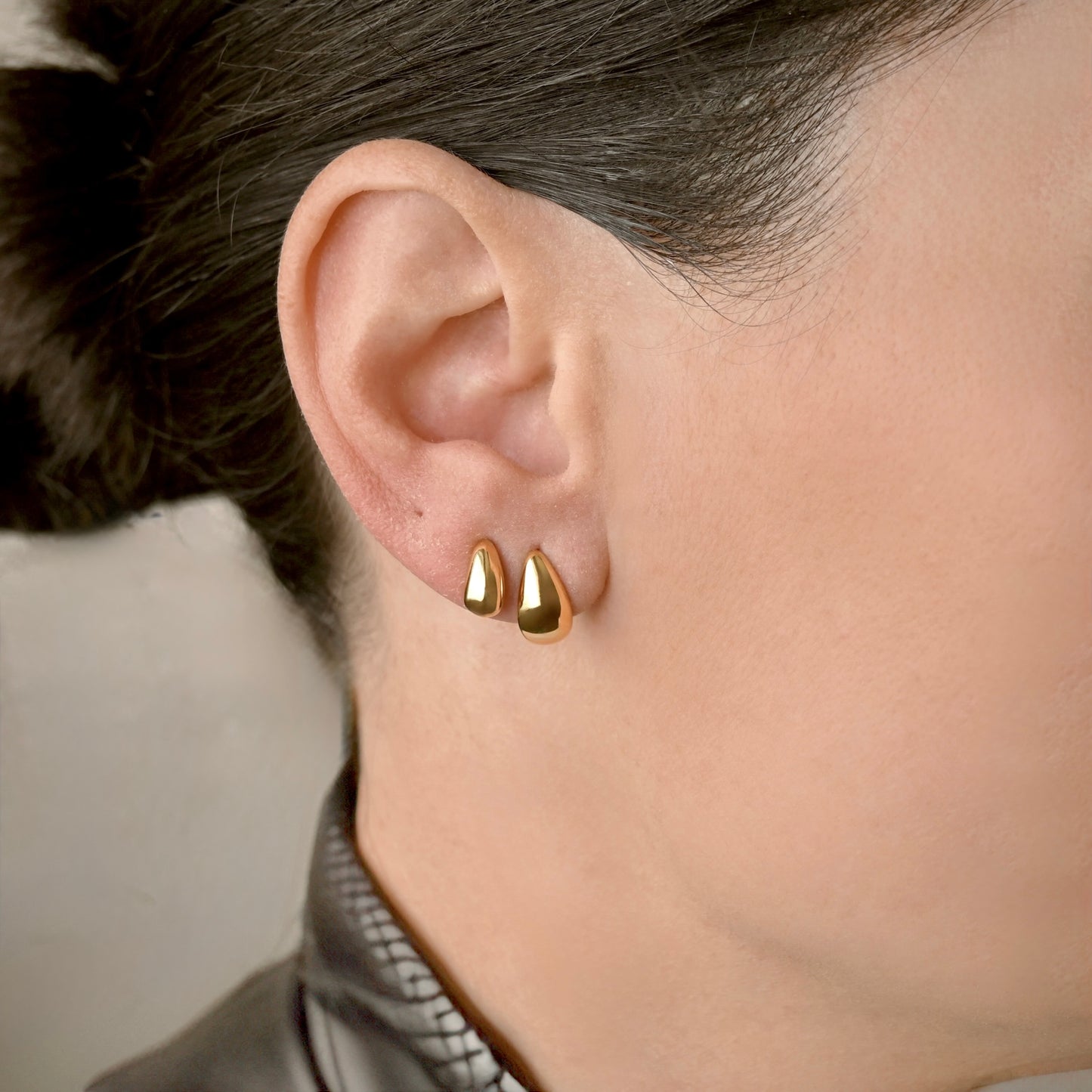 Close-up of a person wearing one small and one medium sized gold dome huggie earrings with a neutral background