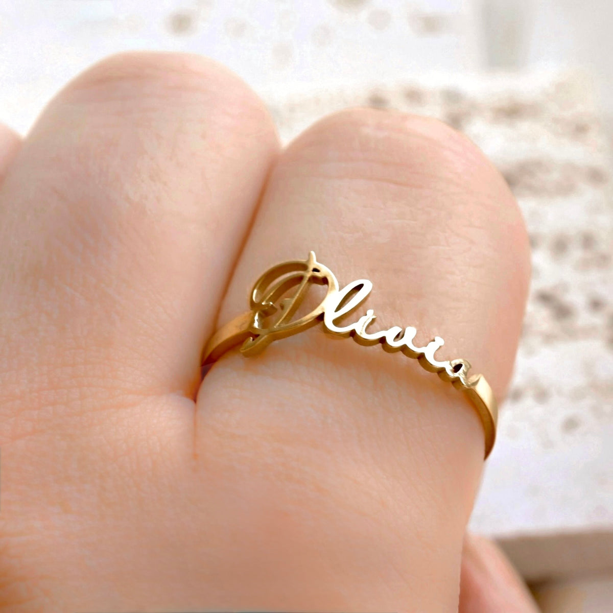 A gold name ring engraved with the name 'Olivia' in cursive script, shown worn on a finger.