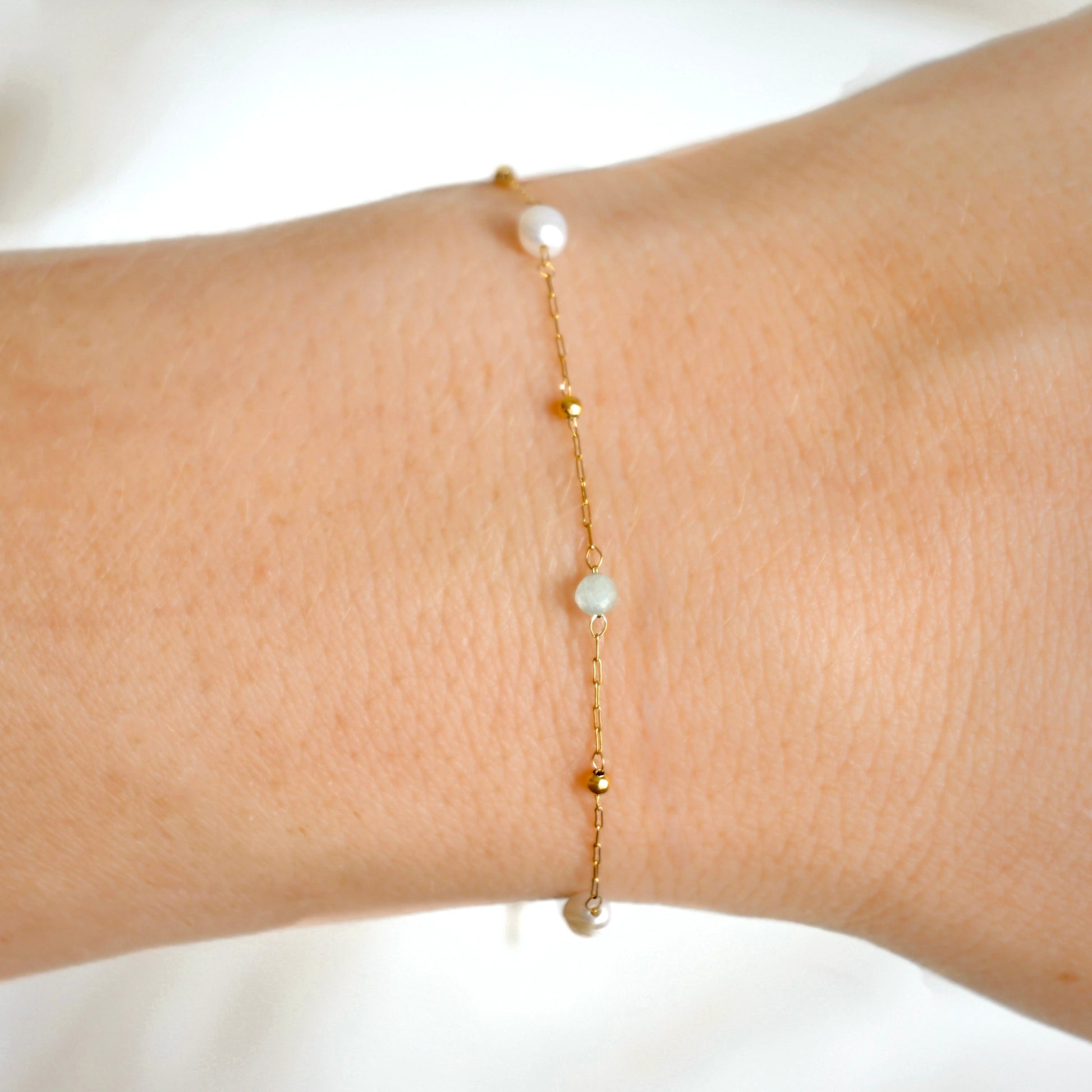 A delicate gold chain bracelet with small gold beads and two white pearls and gold sage beads , shown worn on a person's wrist against a soft white backg background