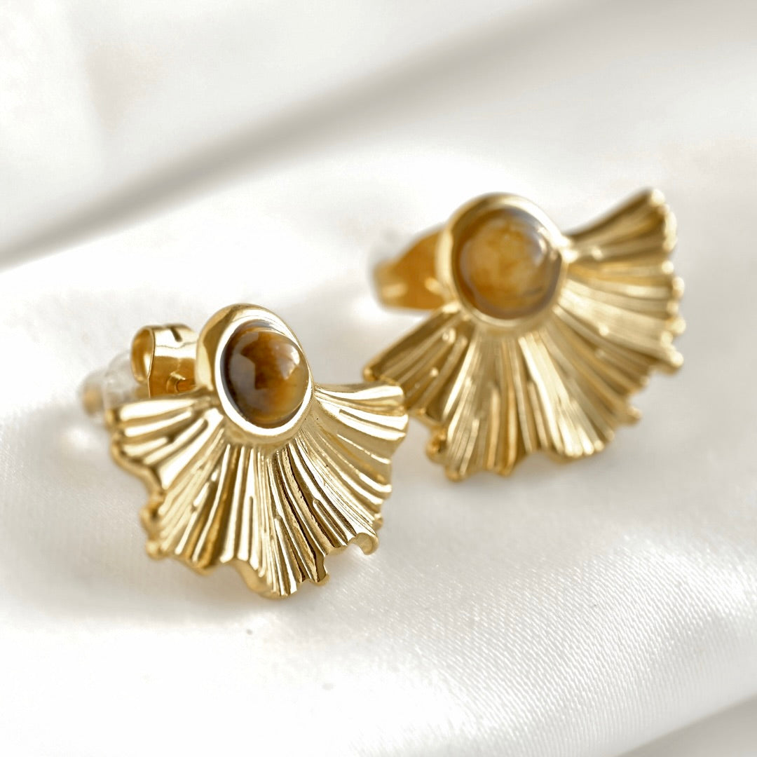 A pair of gold fan-shaped stud earrings with oval brown tiger's eye stones, displayed on a white satin fabric background.