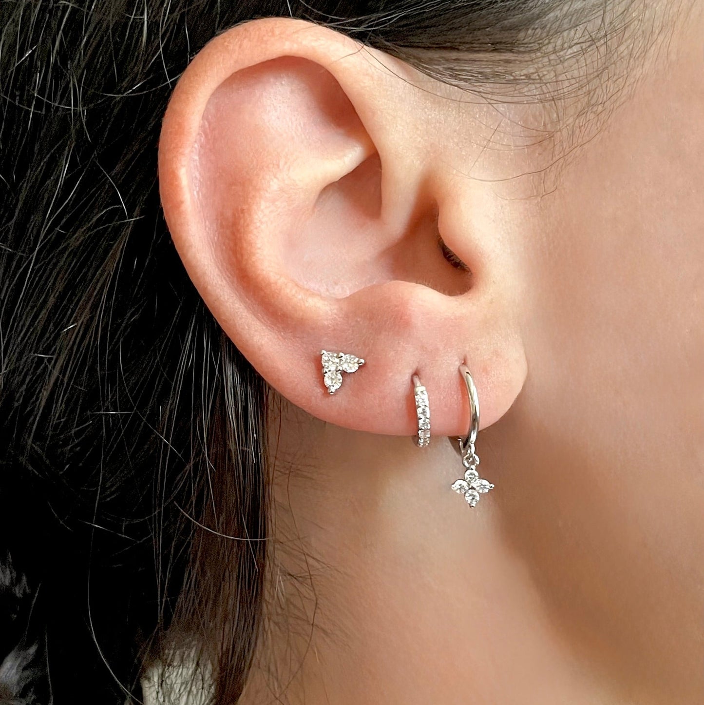 Close-up of an ear wearing silver earrings, a 4-leaf clover dangle hoops, pave huggie earrings, and 3-leaf clover stud earrings with a blurred background