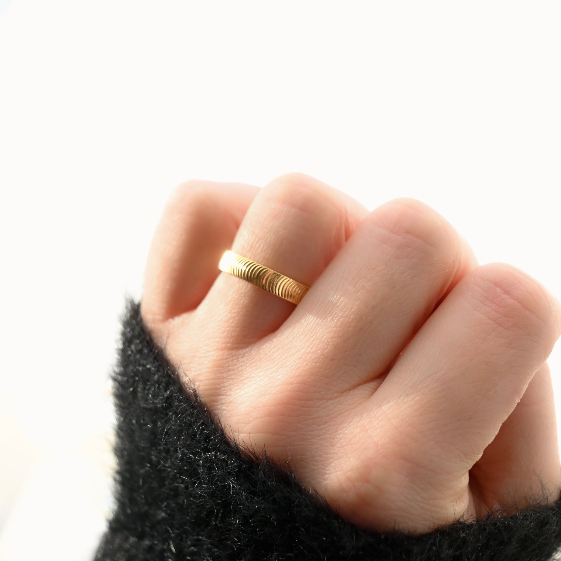 A close-up of a hand wearing a thin, textured gold band ring, set against a bright white background with a black fuzzy sleeve visible at the bottom.