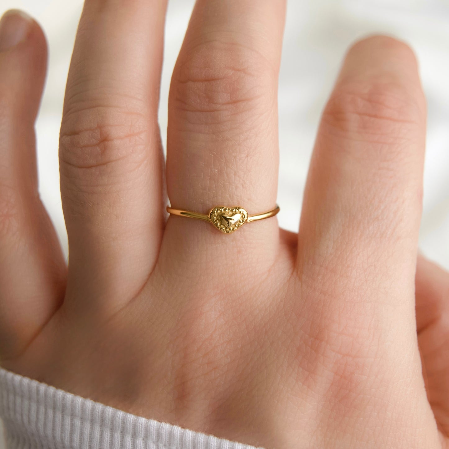Close up view of a delicate gold ring with a small heart-shaped charm, worn on a person's ring finger.