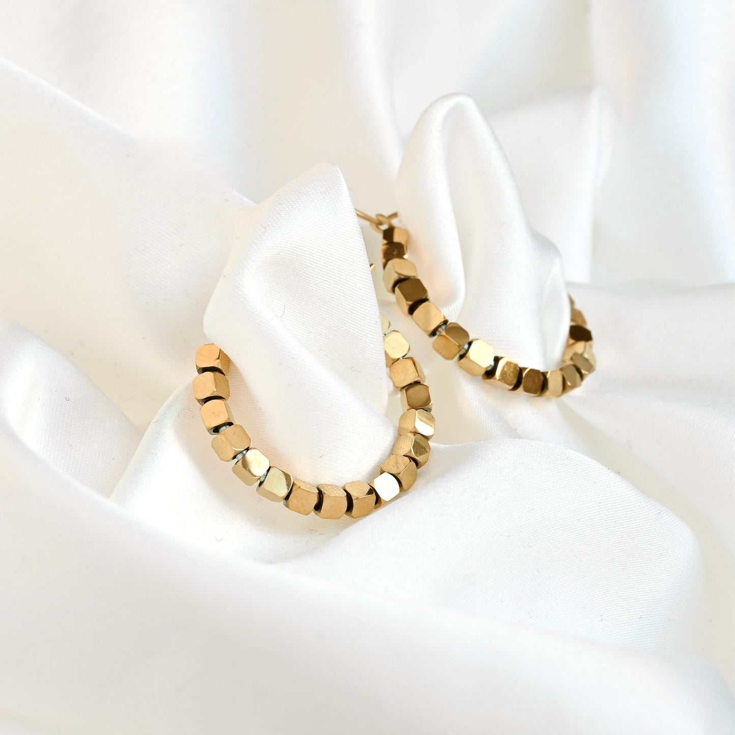 Gold hoop earrings with a faceted, cube-like link design, displayed on a draped white satin fabric background.