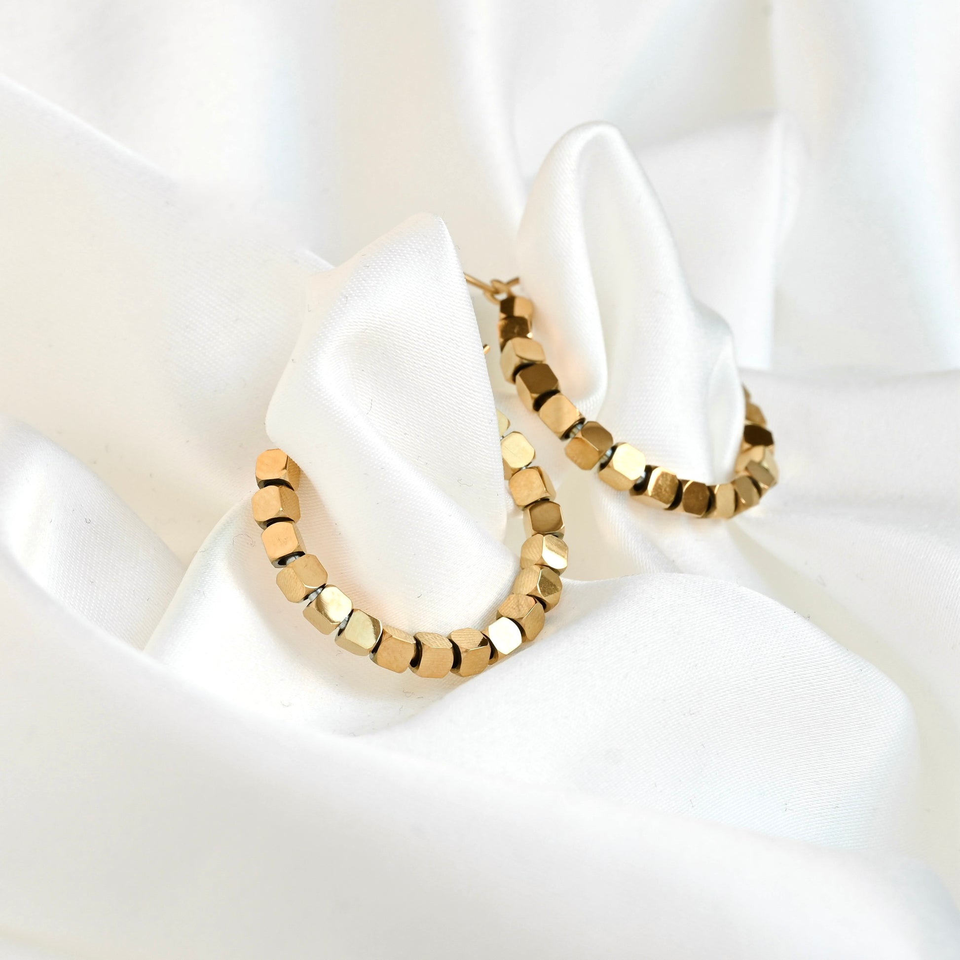 Gold hoop earrings with a faceted, cube-like link design, displayed on a draped white satin fabric background.