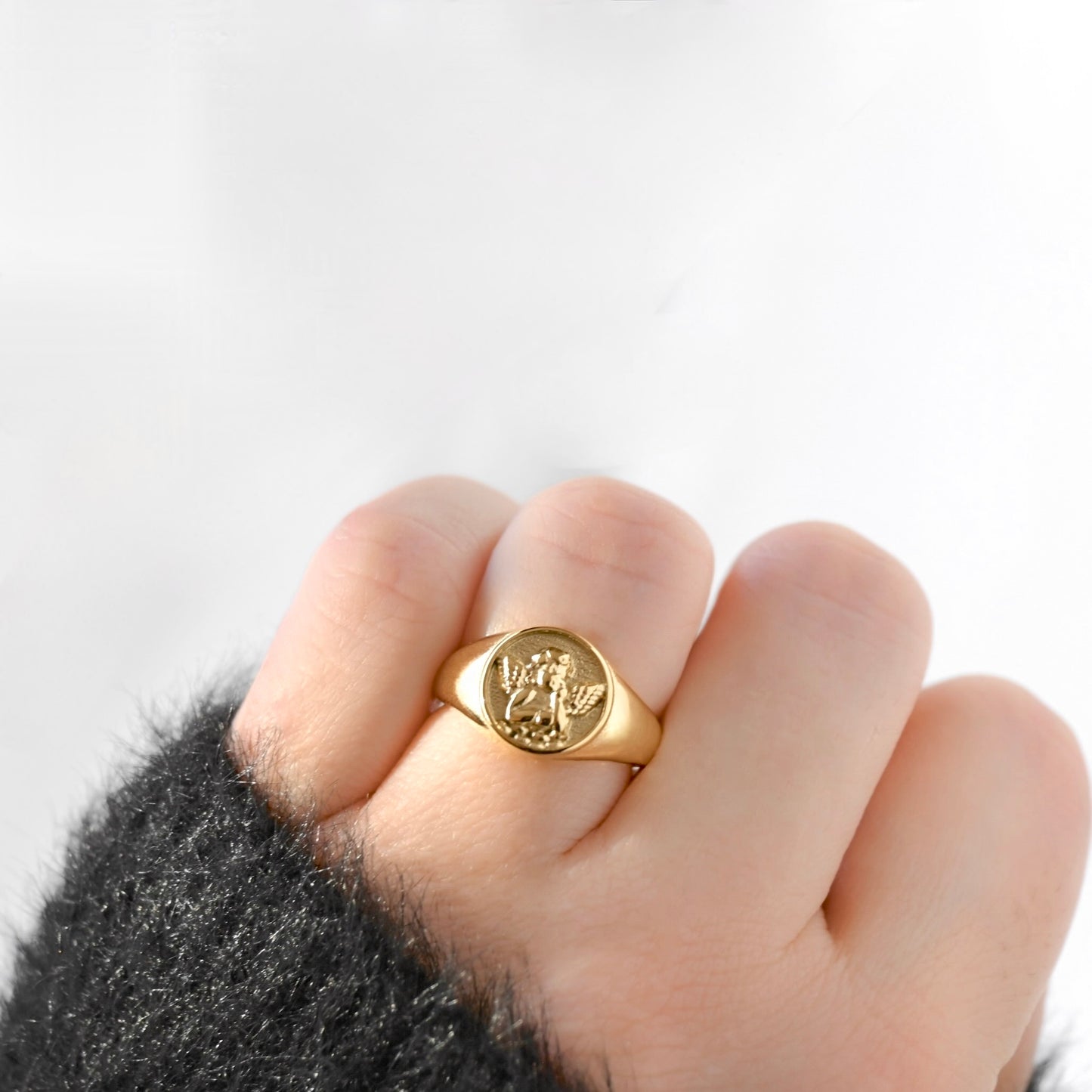 A gold signet ring with an engraved angel figure, worn on a person's finger with a dark fuzzy sleeve visible at the wrist, against a plain white background.