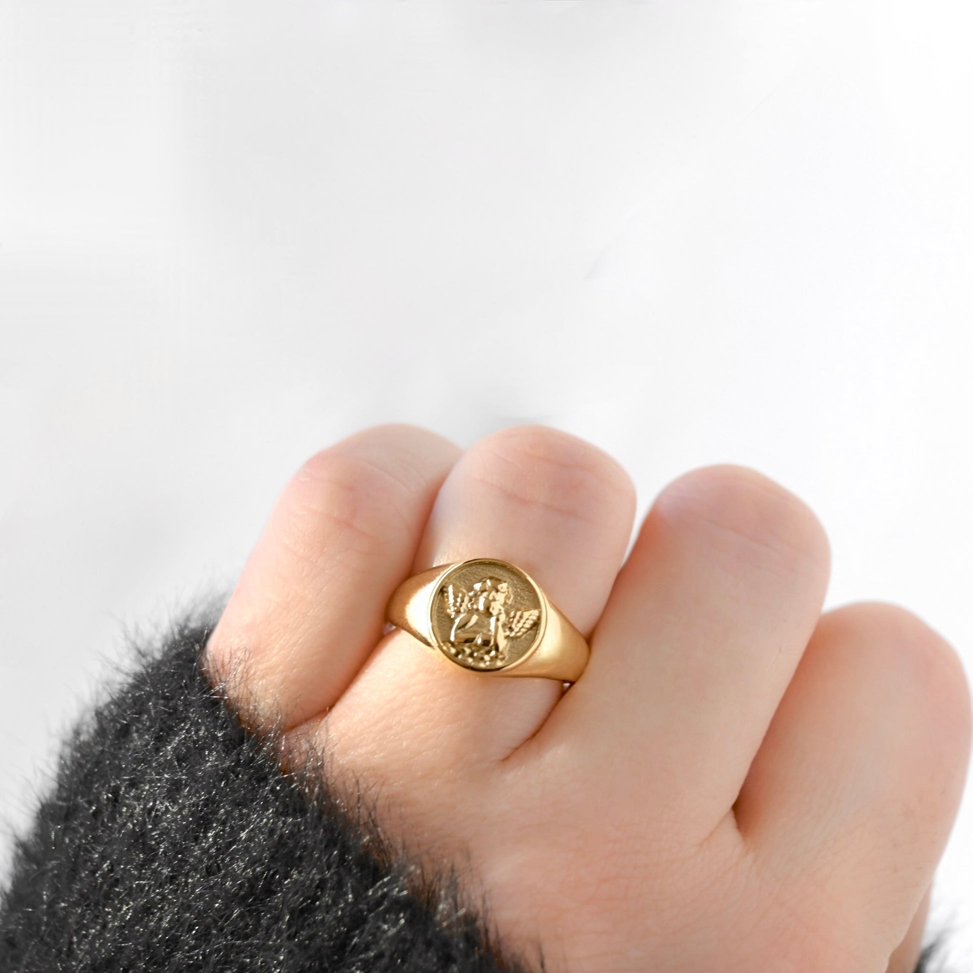 A gold signet ring with an engraved angel figure, worn on a person's finger with a dark fuzzy sleeve visible at the wrist, against a plain white background.