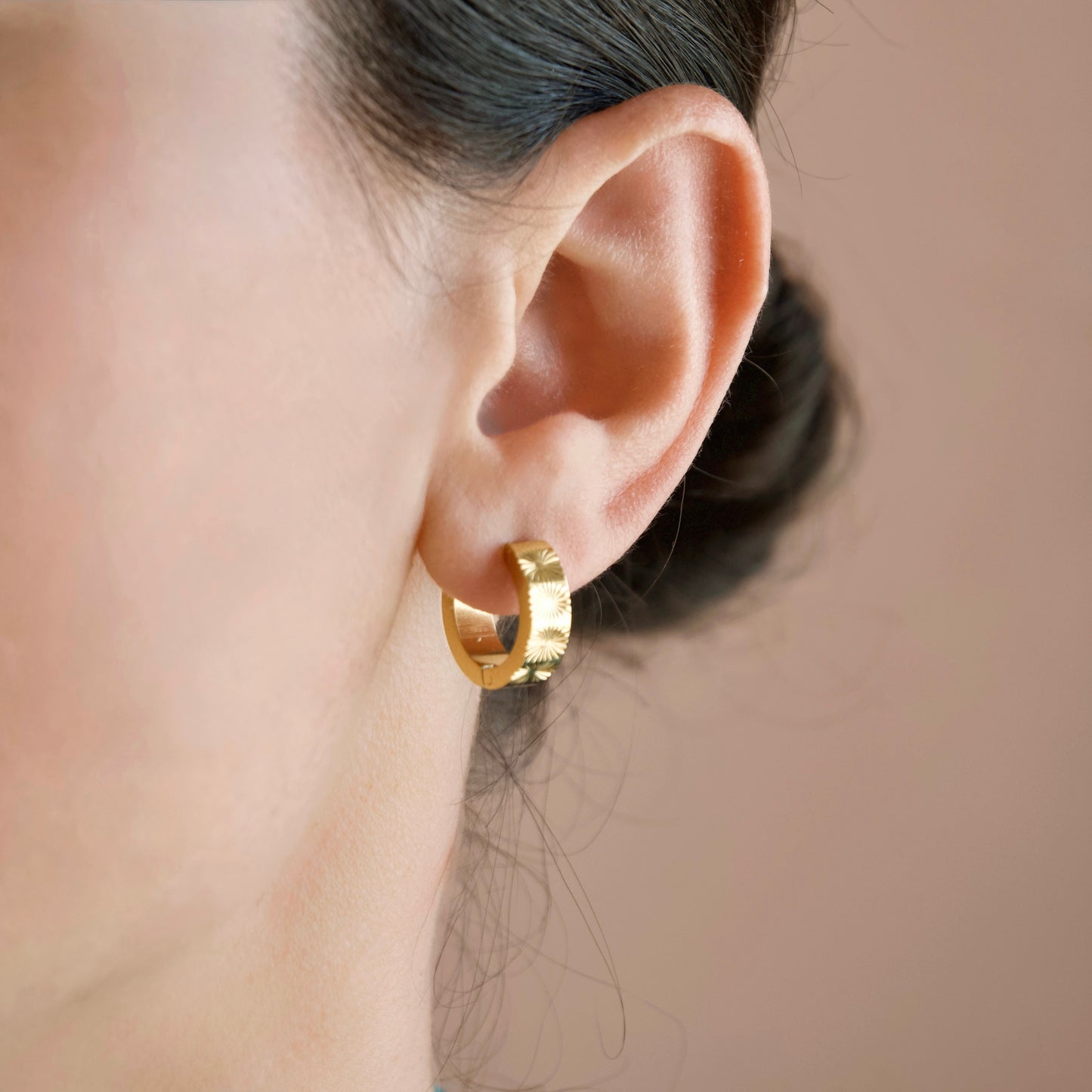 A close-up of a person's ear wearing a small thick gold huggie hoop earring against a soft beige background.