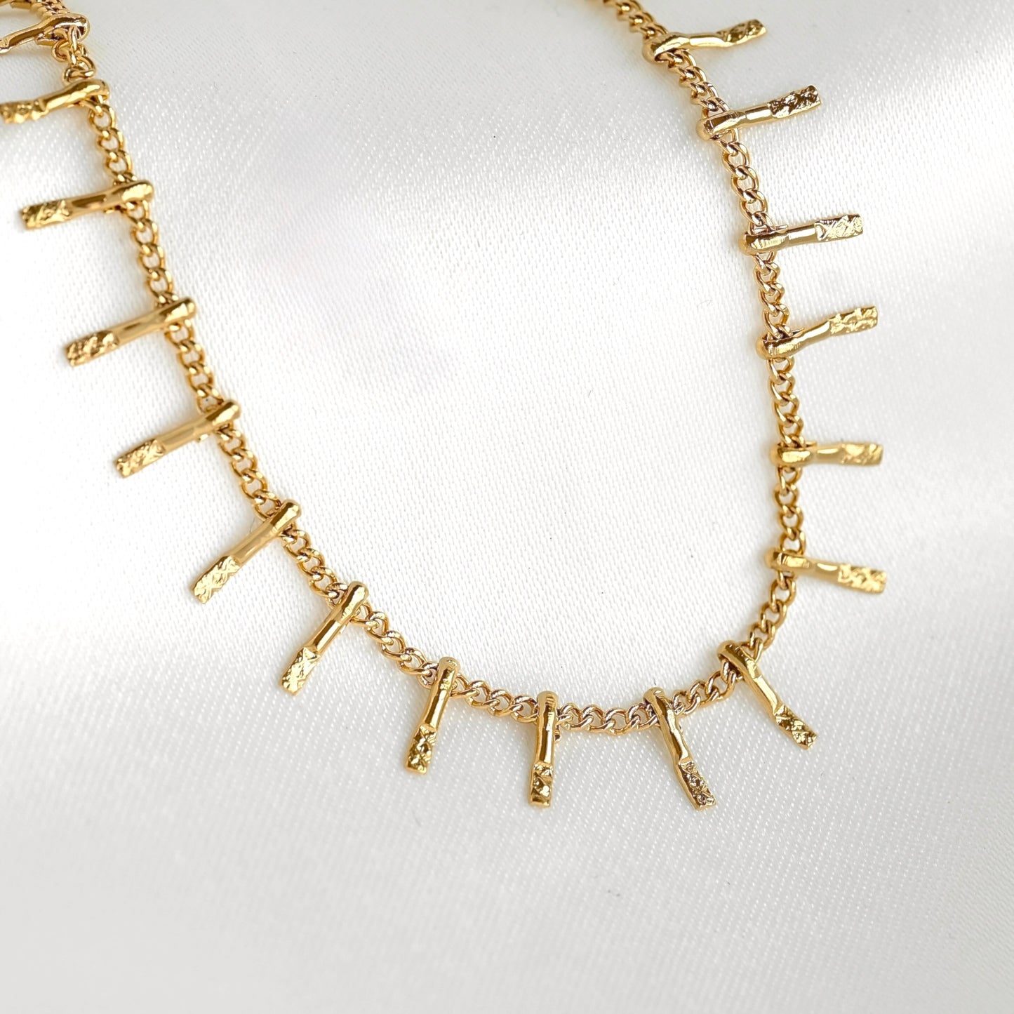 A gold chain necklace featuring textured, branch-like metallic pendants arranged in a line, displayed on a soft white fabric background.