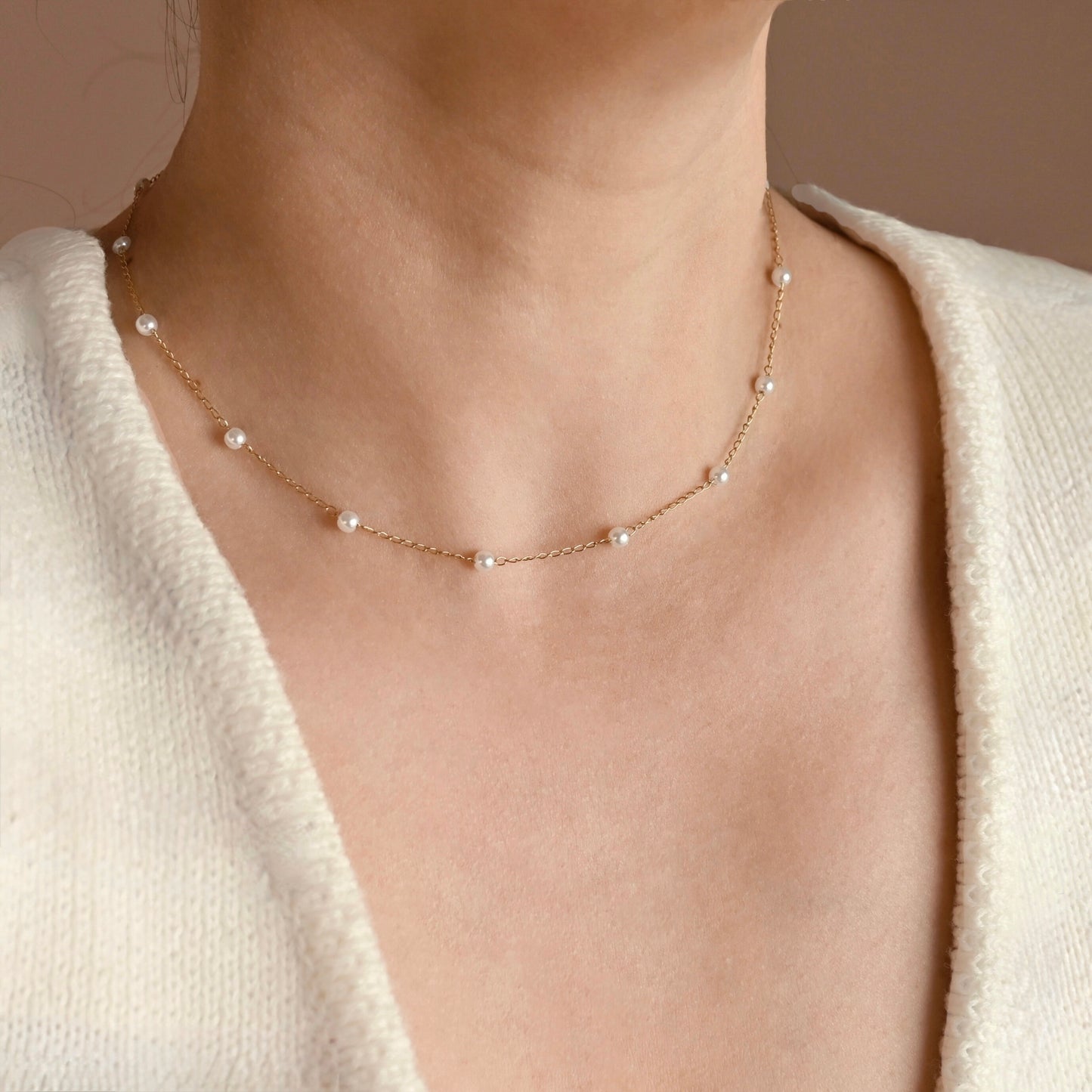 A delicate gold chain necklace with small white pearls spaced along the chain, worn on a model's neck over a cream-colored knit top.