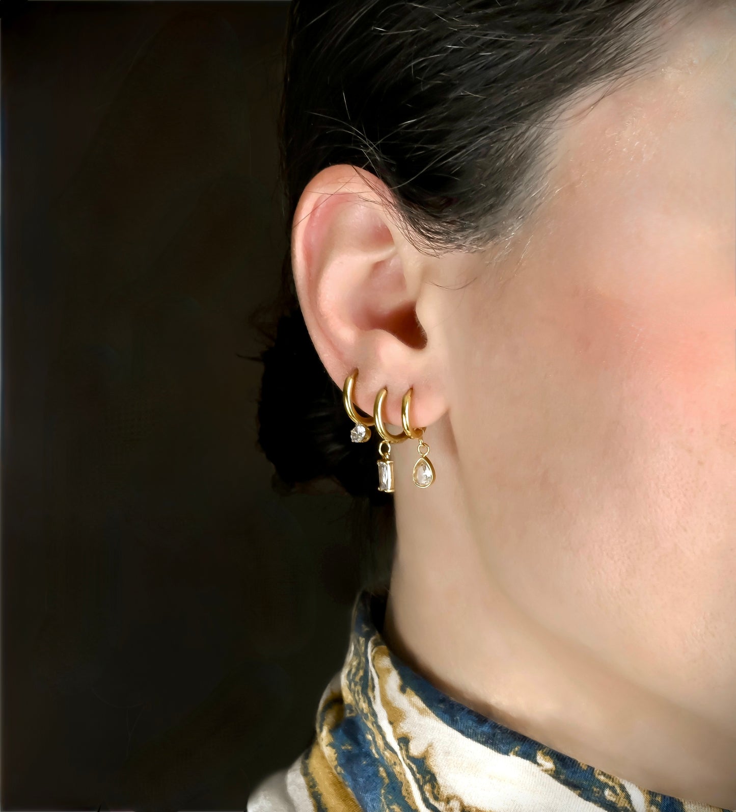 Close-up of an ear wearing gold hoop earrings with small gemstones against a dark background.