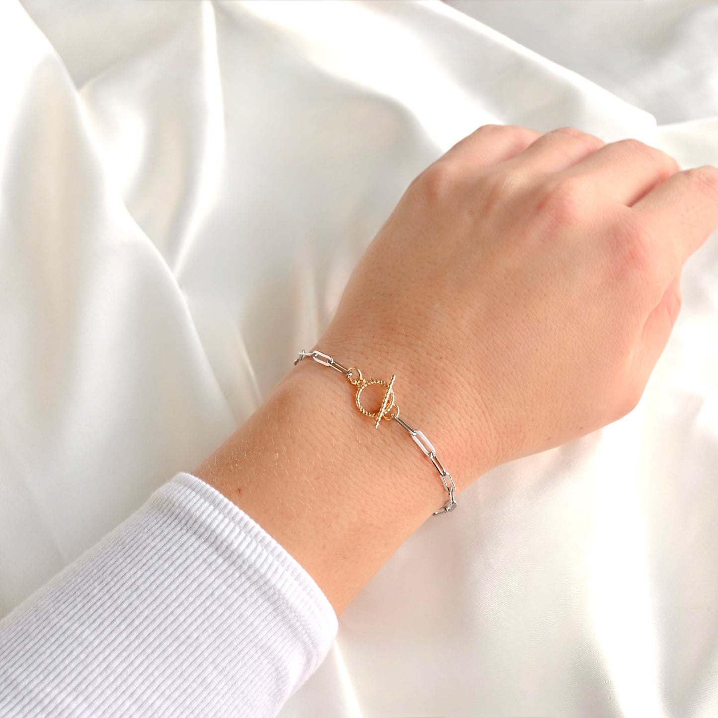 A delicate two-tone bracelet with a gold ring and toggle clasp on a silver chain, shown worn on a person's wrist against a soft white fabric background.