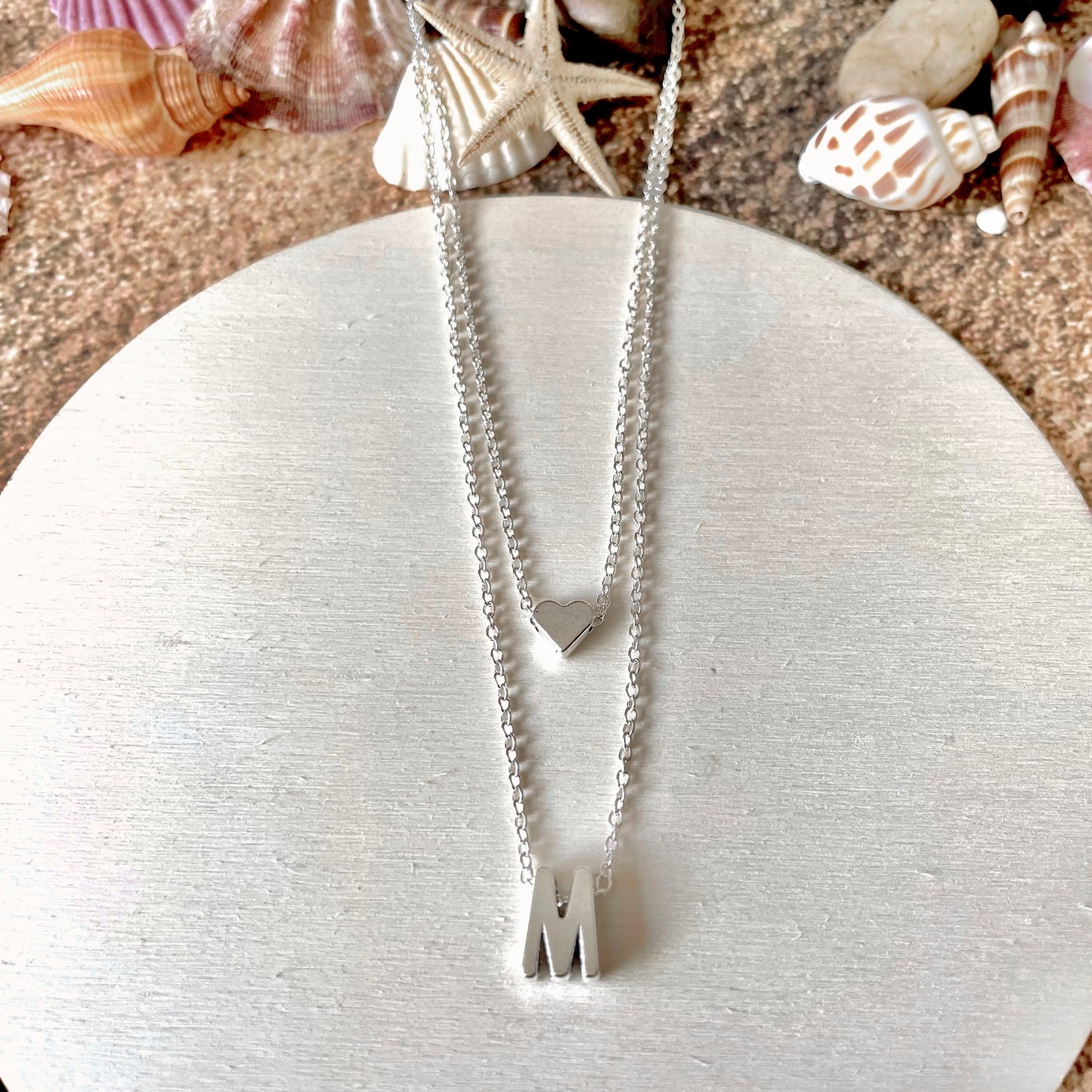 Silver necklace with letter 'M' pendant and heart charm on a white wooden plate with seashells in the background