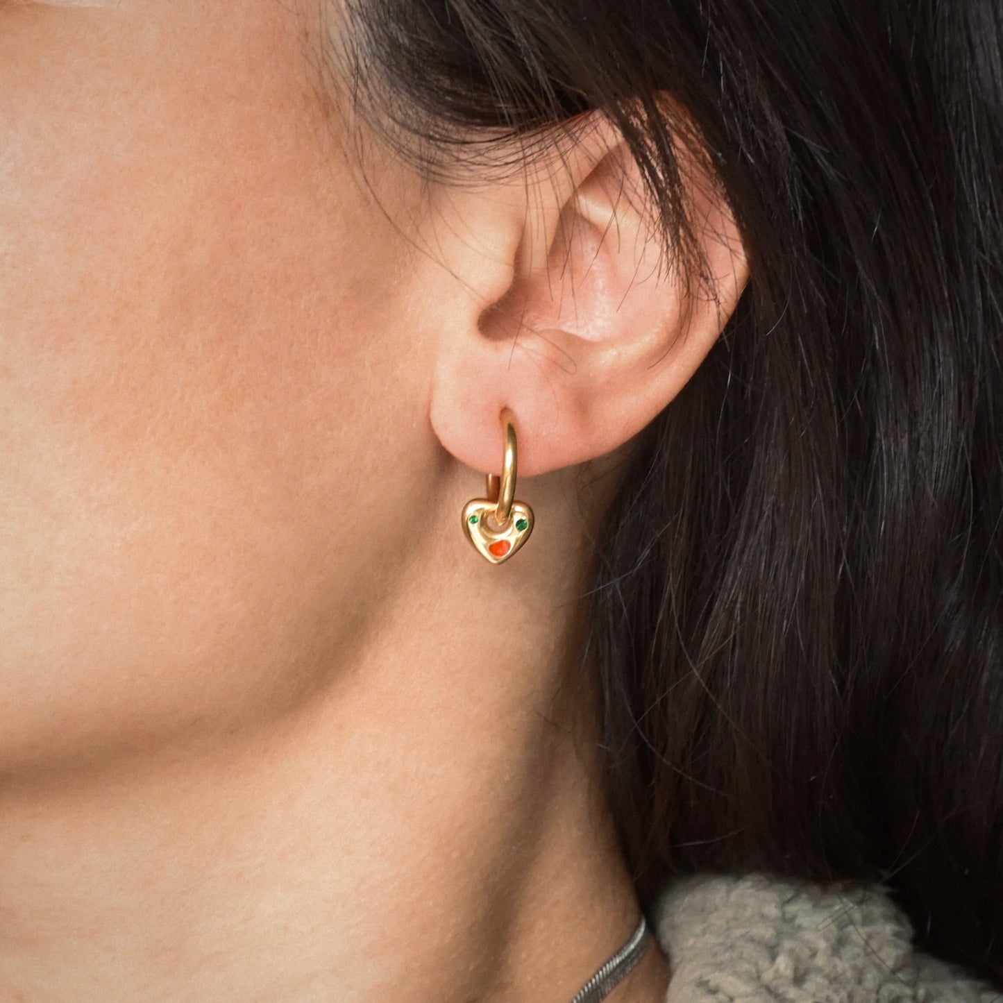 Close-up of a person wearing a gold heart dangle hoops with green gemstones.