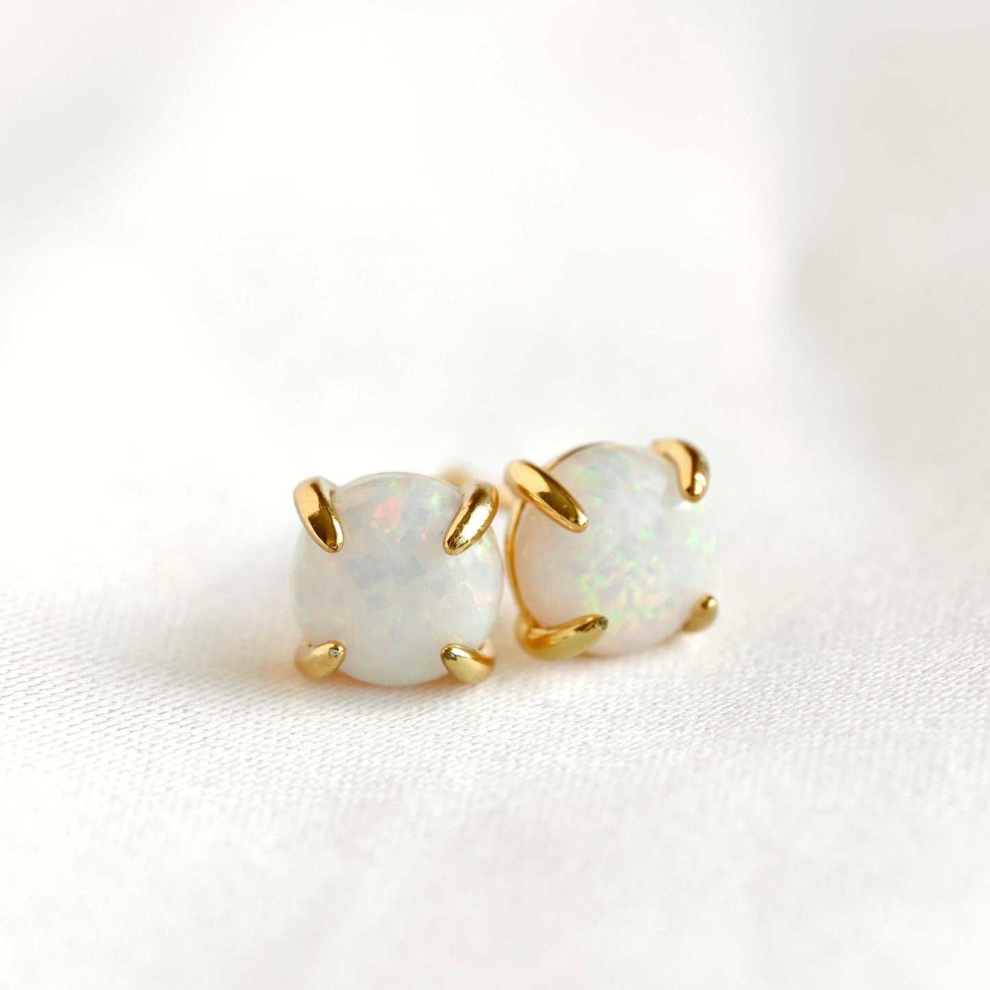Front view of a pair of gold stud earrings with oval white opal gemstones, set in a four-prong setting on a soft white fabric background.