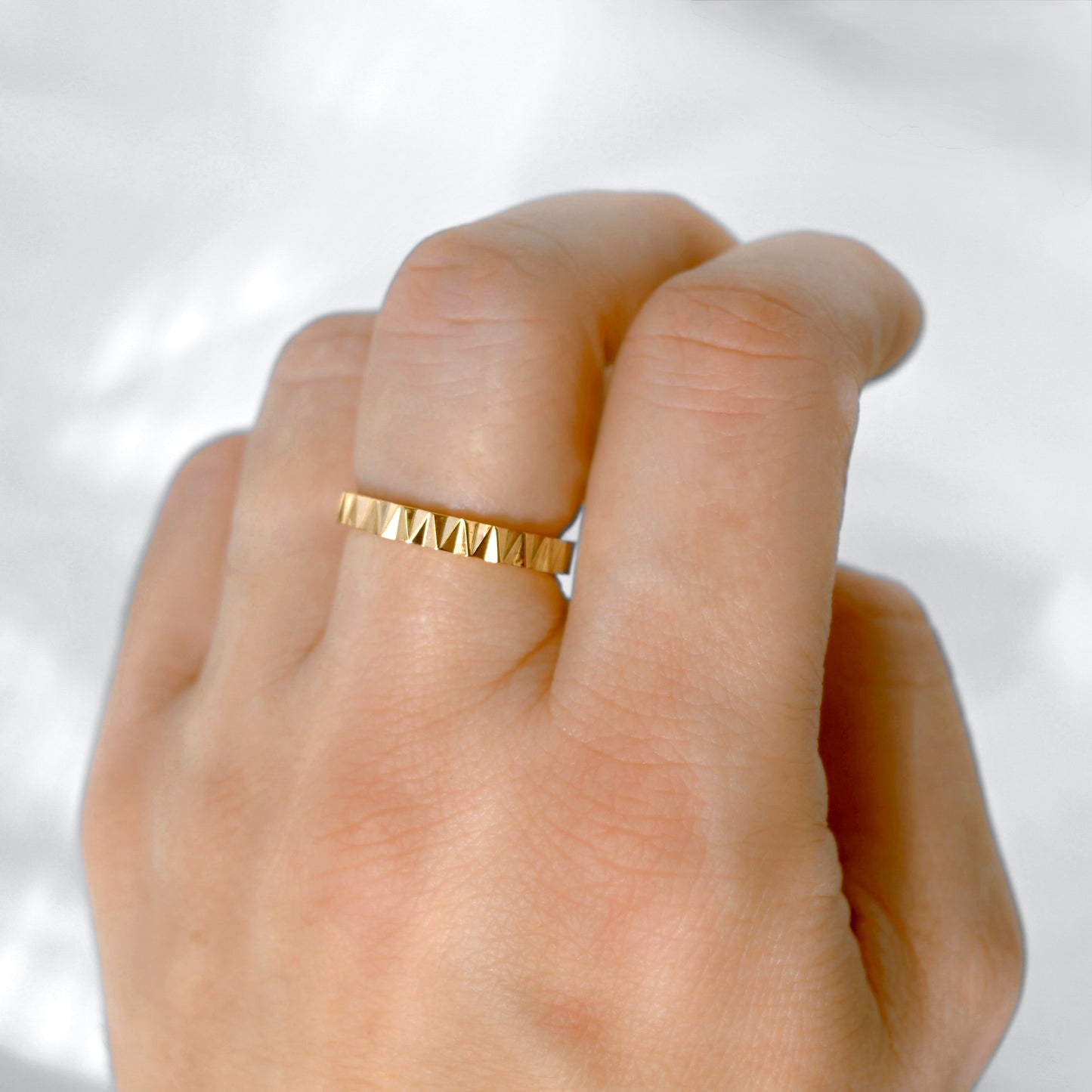 A close-up of a hand wearing a thin gold band ring with a repeating geometric triangular pattern, set against a soft, light gray background.