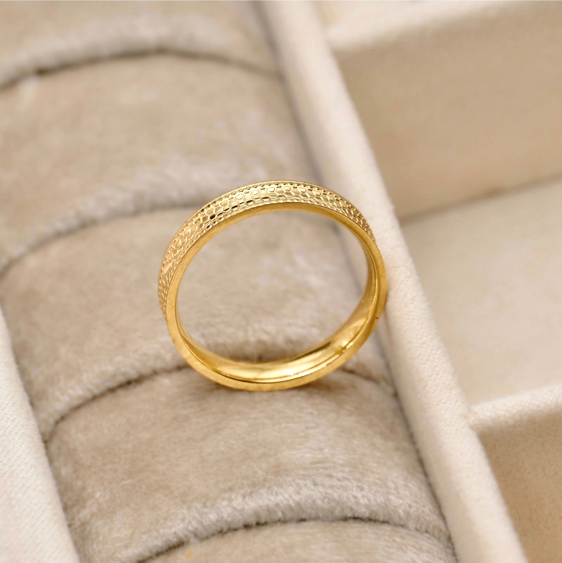 A gold band ring with a textured, hammered finish, displayed on a beige velvet ring box.