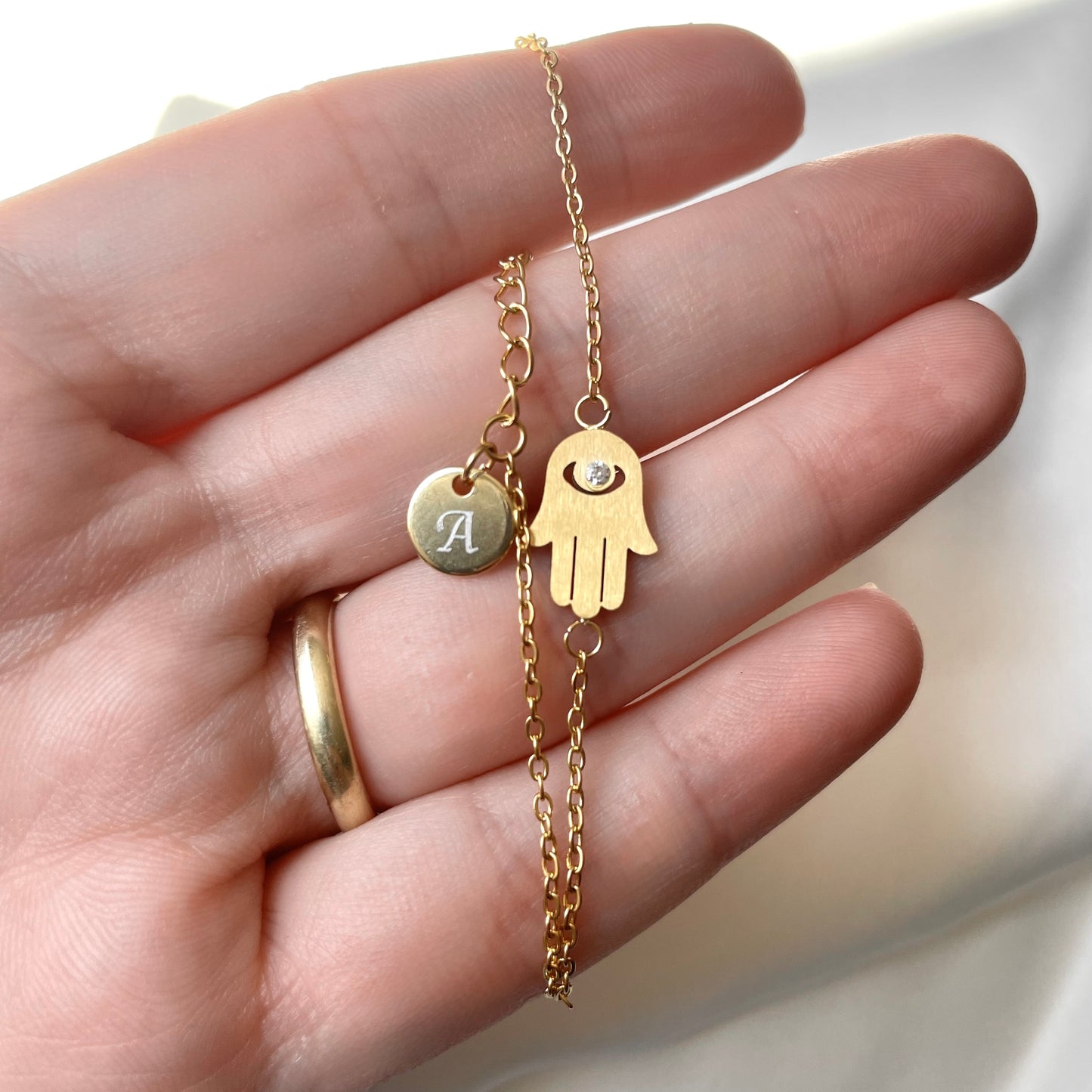 Gold bracelet with a hand-shaped charm and an initial tag at the clasp against a neutral background