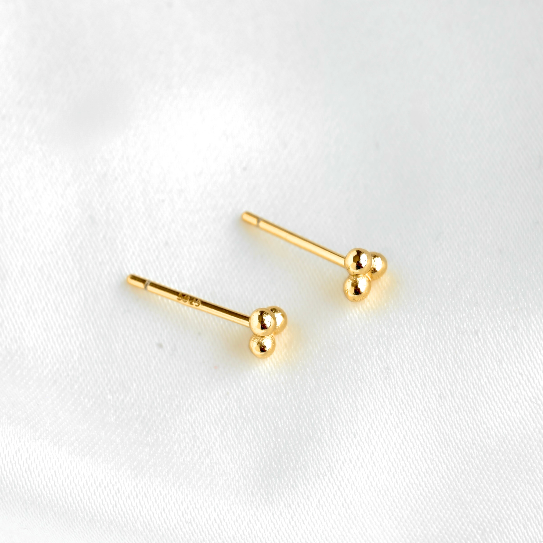 A pair of gold stud earrings with small spherical bead details at the end, displayed on a soft white fabric background.