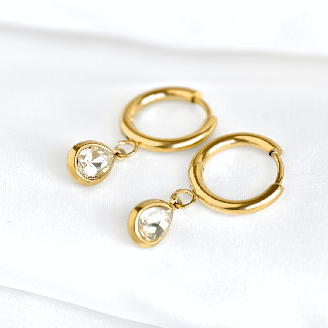 A pair of gold huggie hoop earrings with dangling clear teardrop diamonds in bezel setting, displayed on a soft white fabric background.