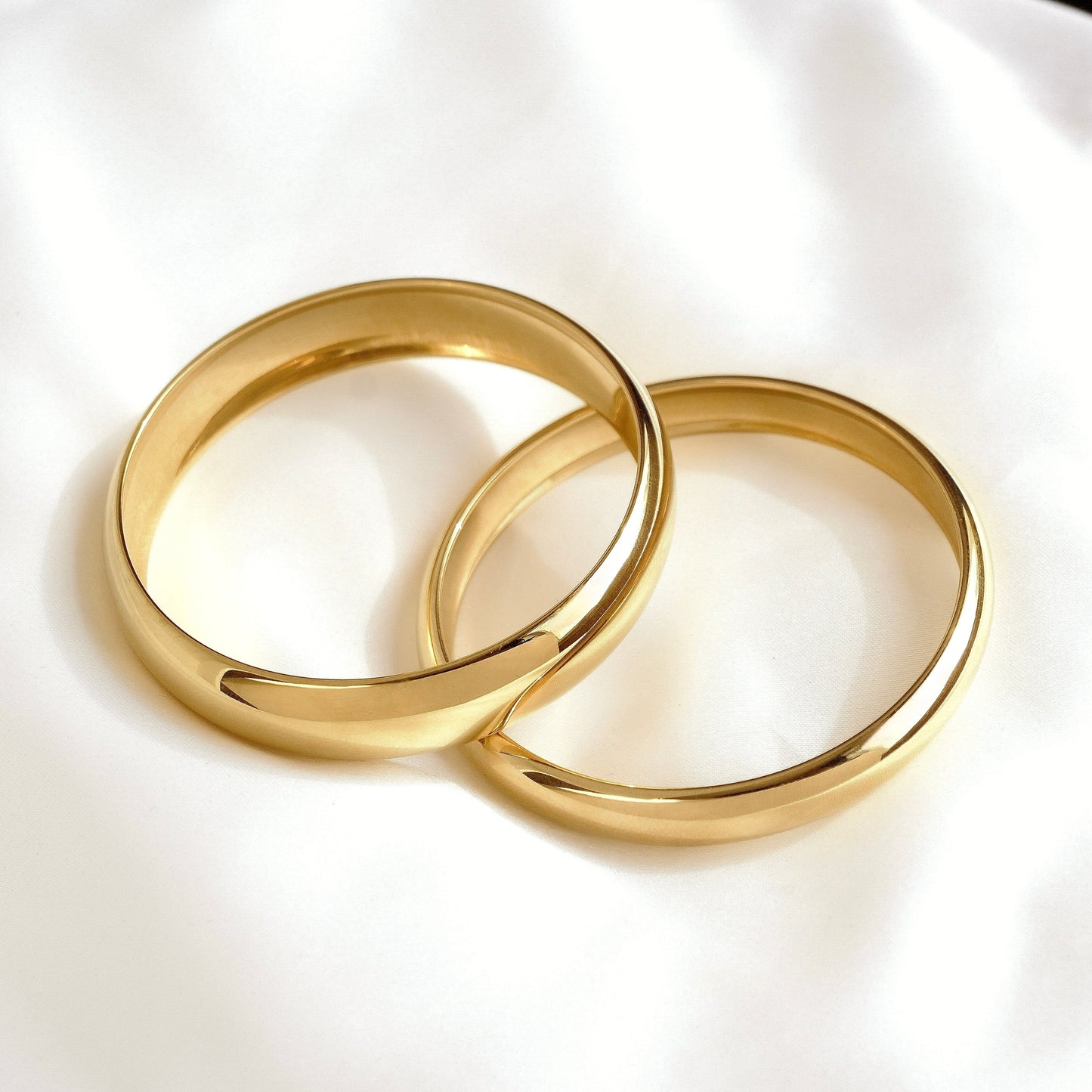 Two polished gold bangle bracelets resting on a soft white fabric background.